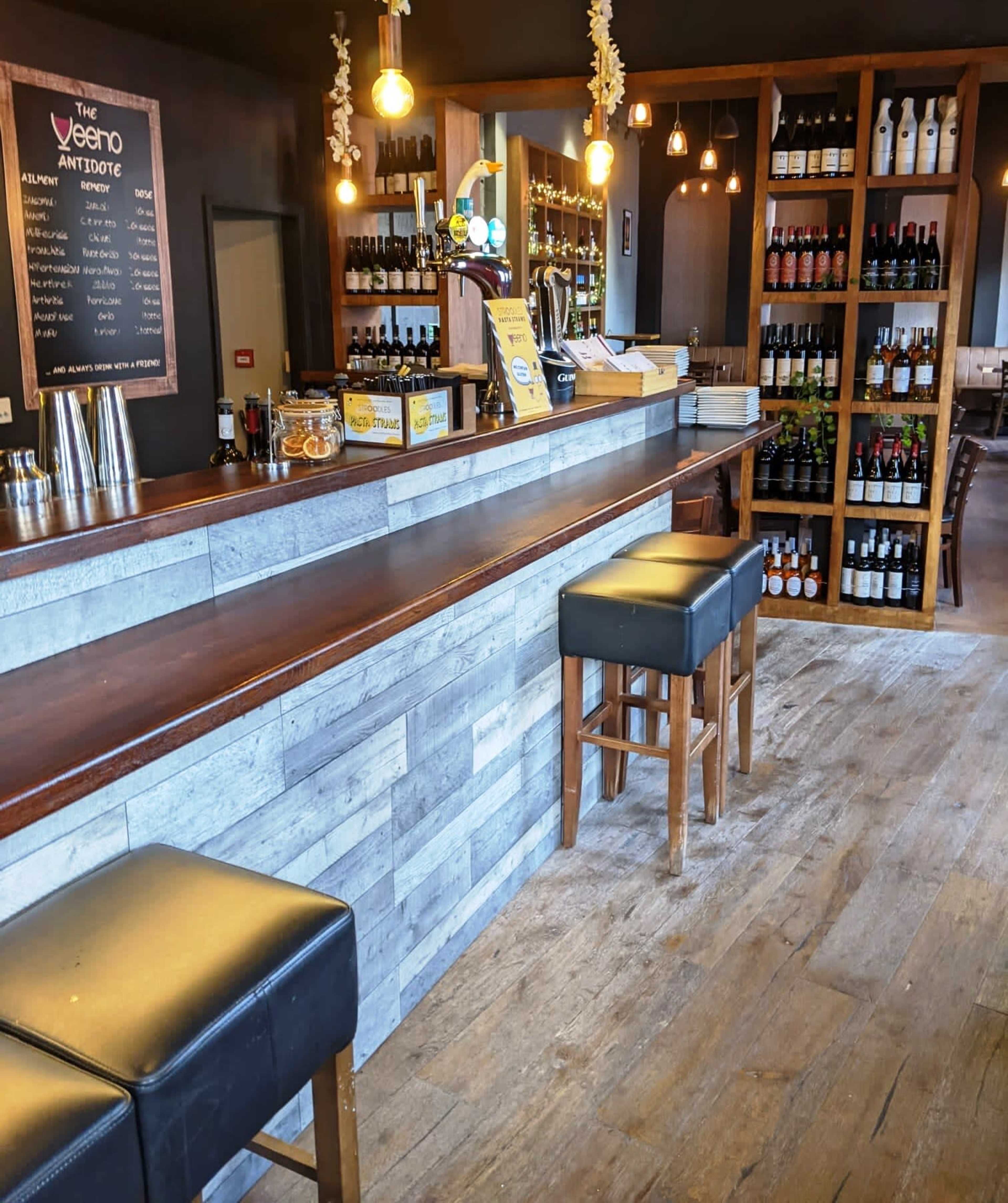 A bar area with a wooden counter, high stools, shelves filled with bottles, and warm lighting.