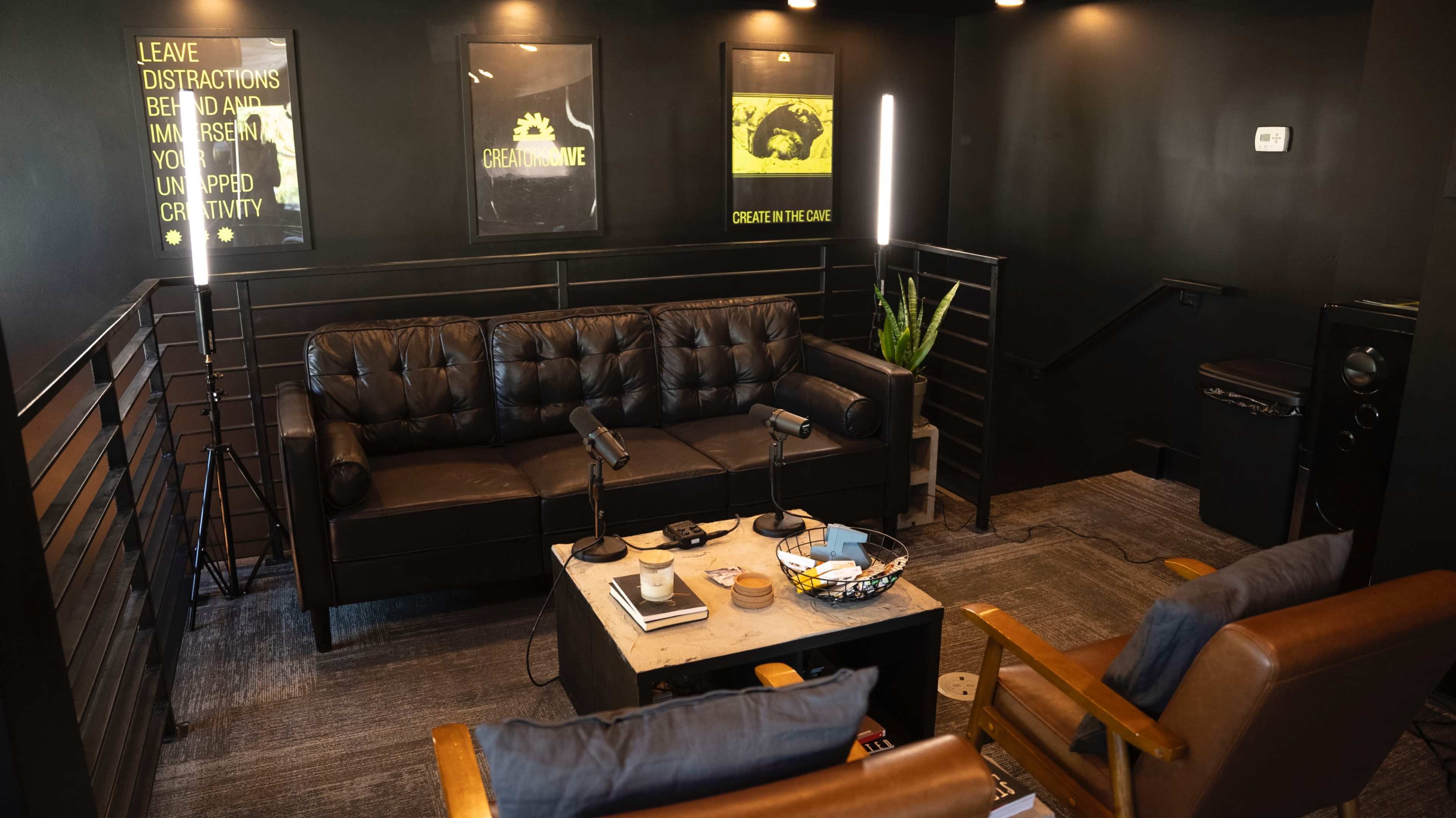 A modern, dimly lit room featuring a black leather sofa, two wooden chairs, a coffee table with various items, and wall art with motivational quotes.