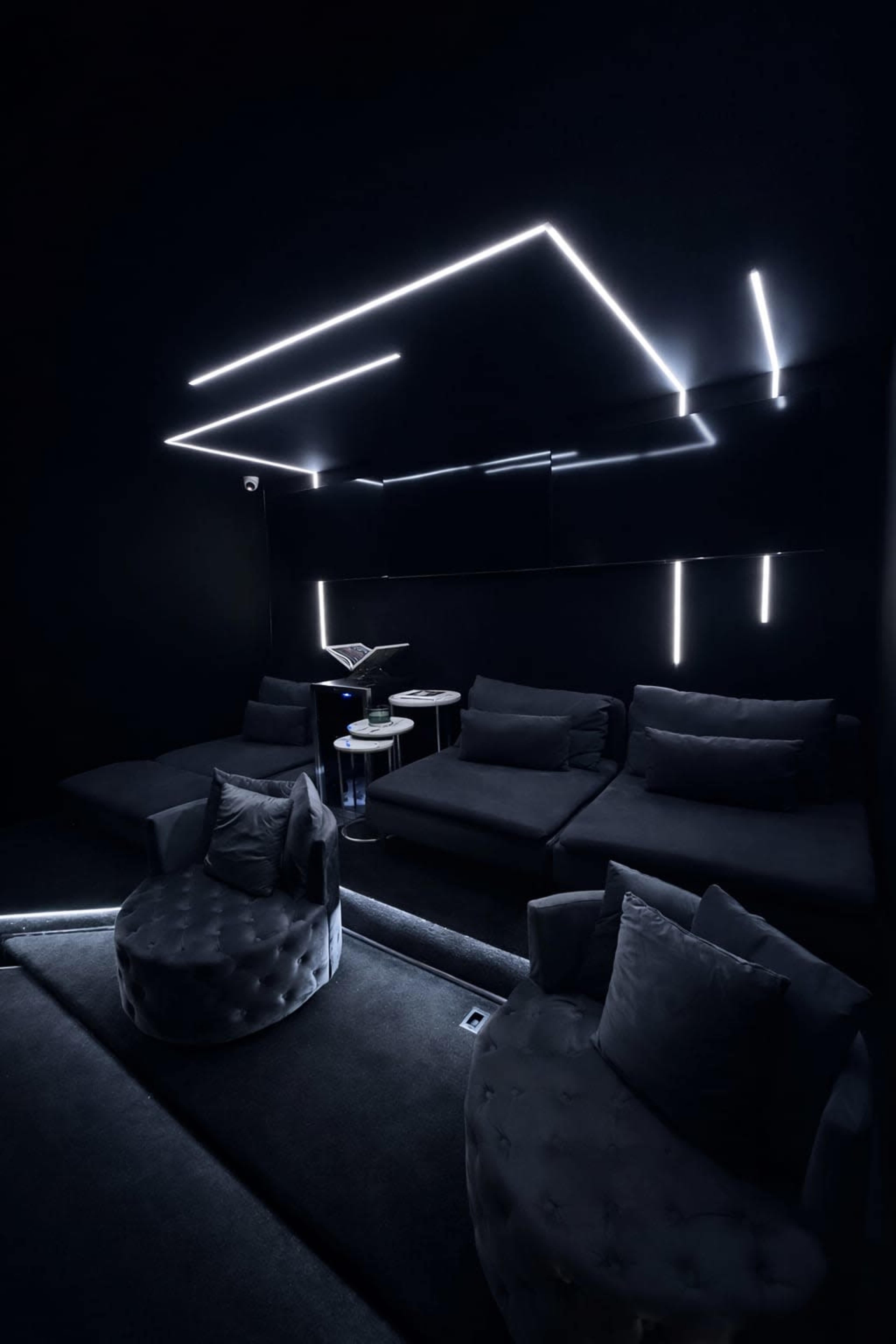 The image shows a dimly lit room featuring dark furniture, modern sofas, and illuminated geometric lines on the walls and ceiling.