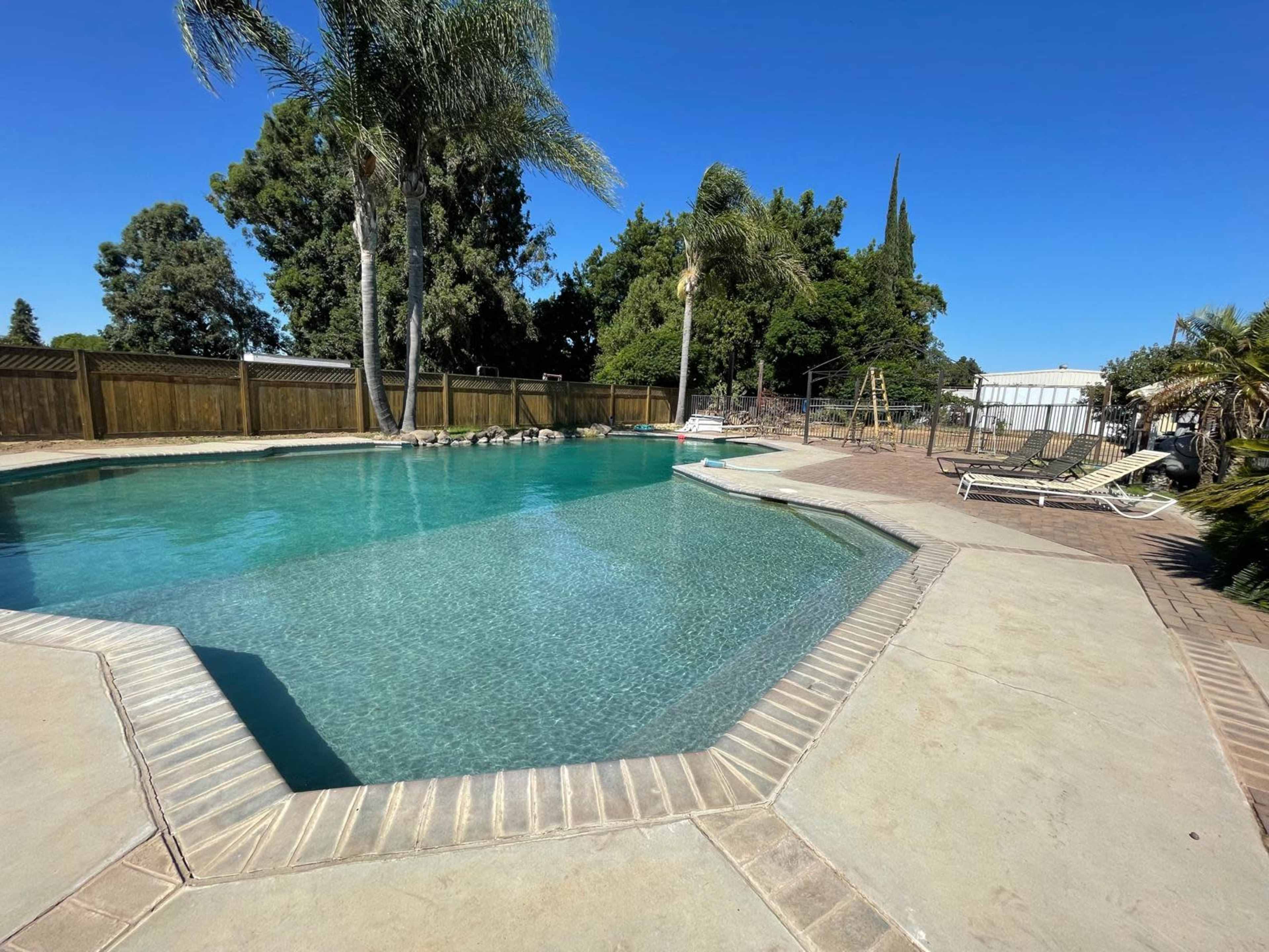 A swimming pool surrounded by a concrete deck, trees, and a playground area in a residential backyard.