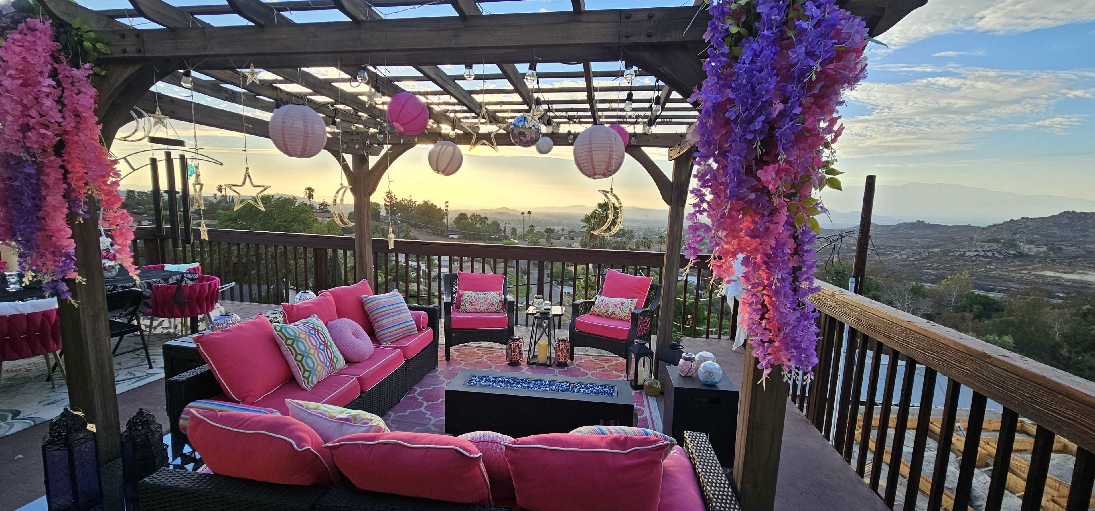 A cozy outdoor seating area is adorned with pink cushions, colorful lanterns, and hanging flowers, overlooking a scenic landscape at sunset.