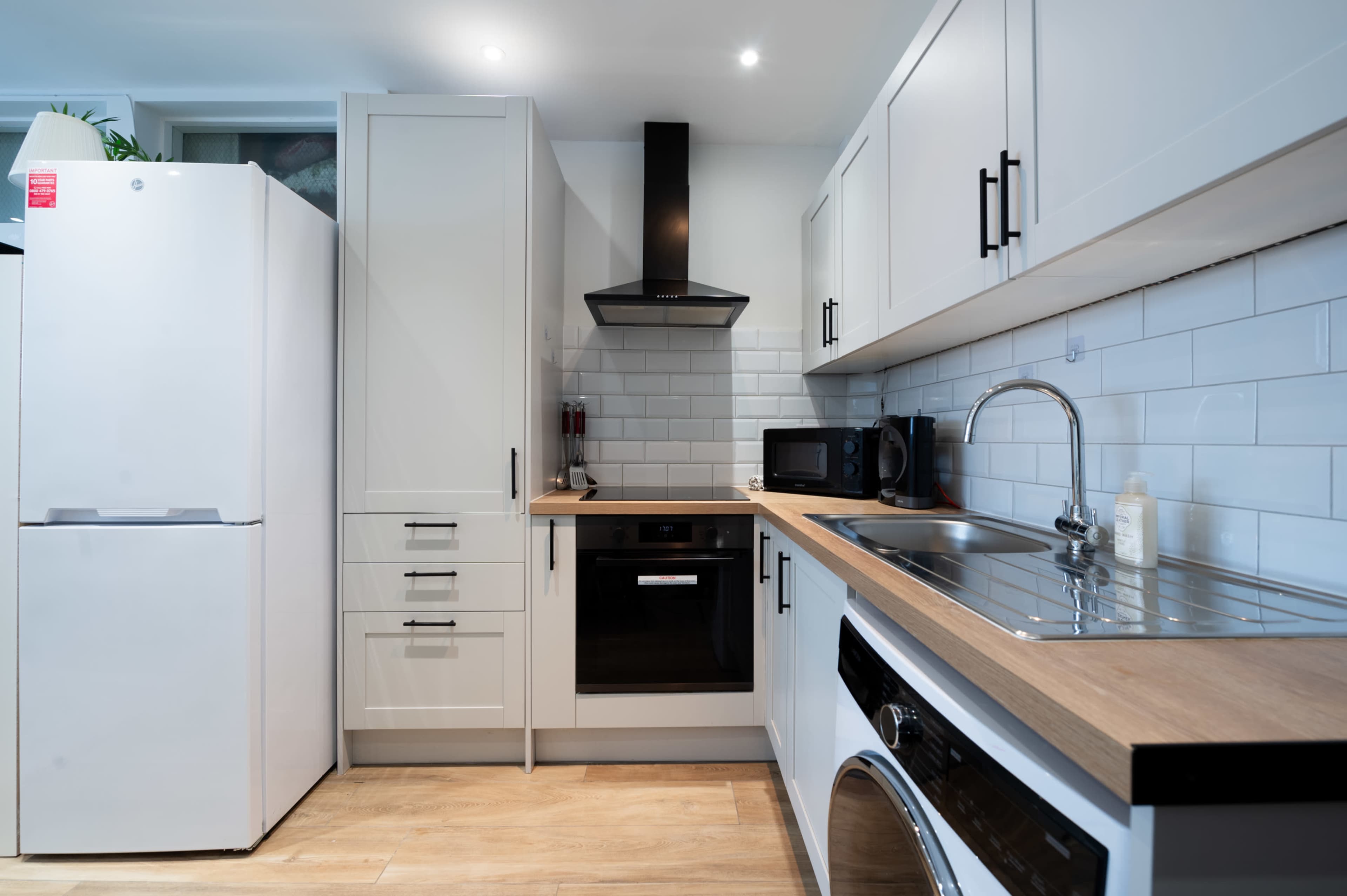 The image shows a modern kitchen with white cabinetry, a stainless steel sink, a black oven, and a refrigerator.