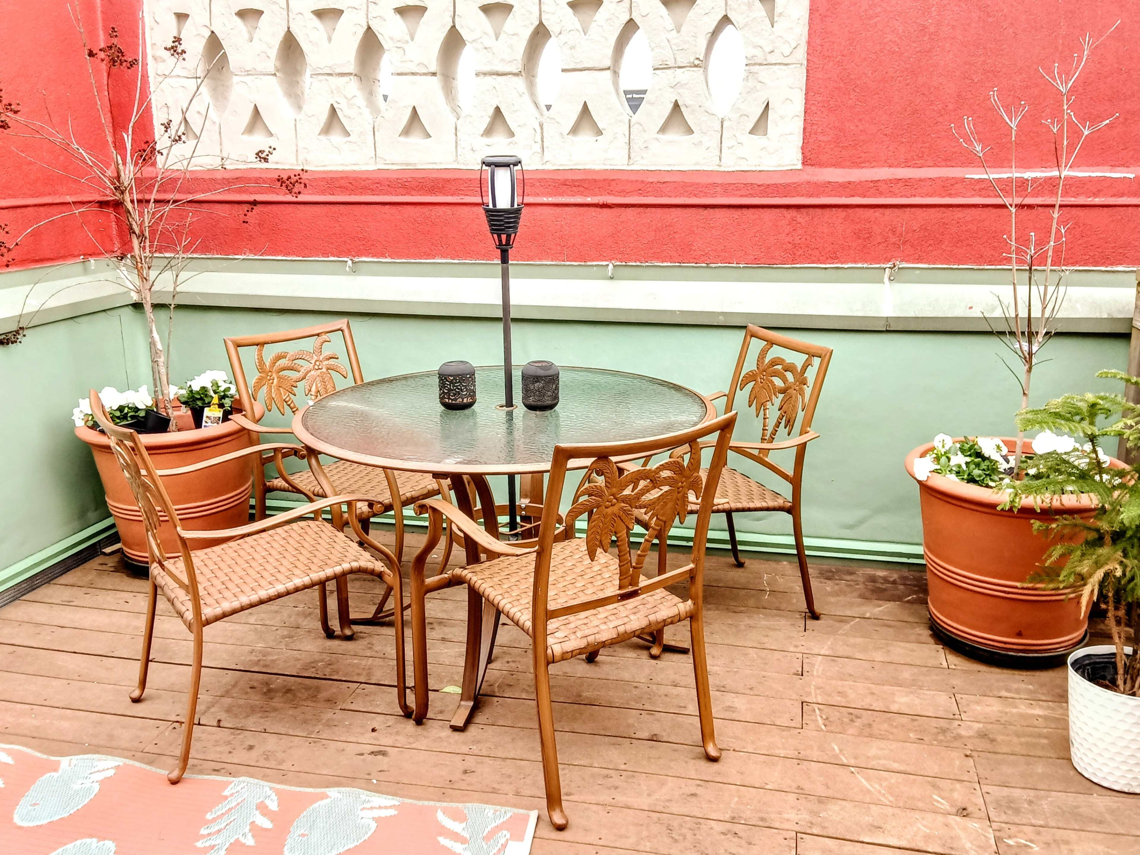 A patio features a circular glass table surrounded by four chairs, with potted plants and a lantern nearby.