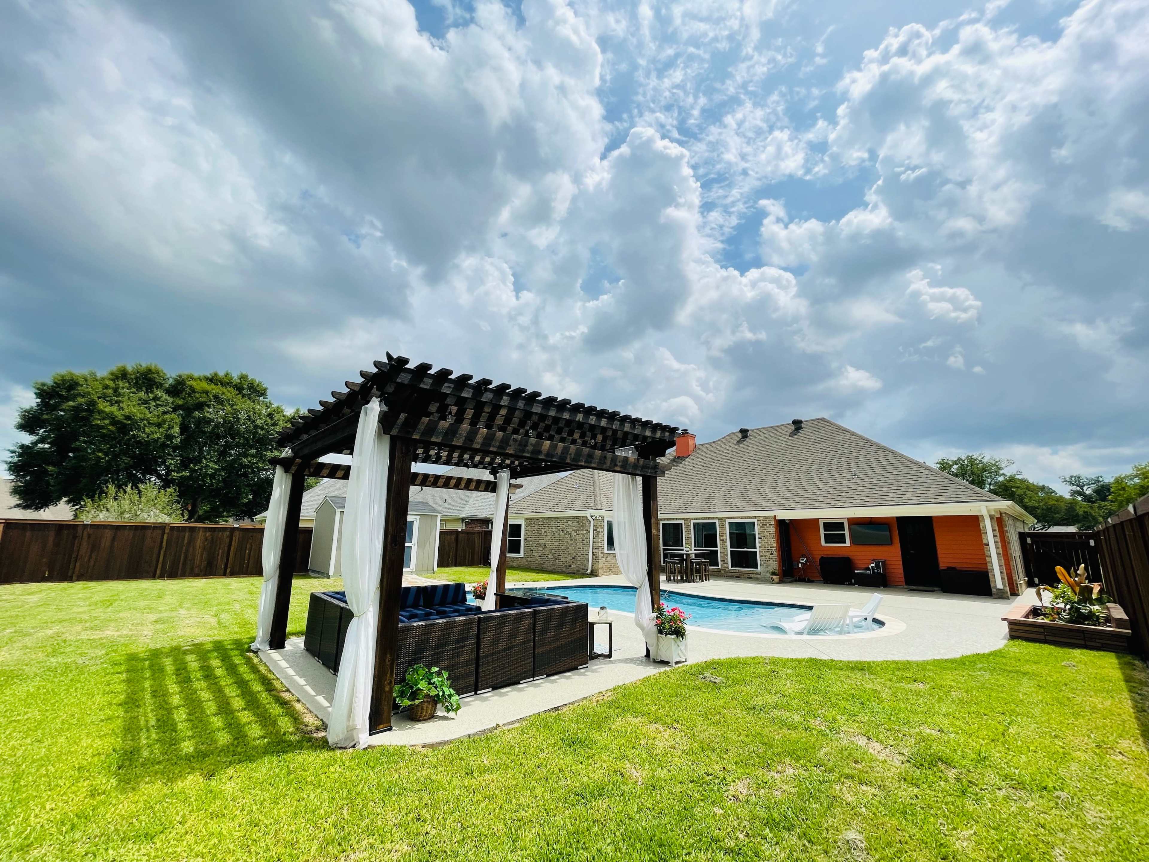 A backyard featuring a pool surrounded by grass, a covered seating area, and a house with a mix of exterior colors.