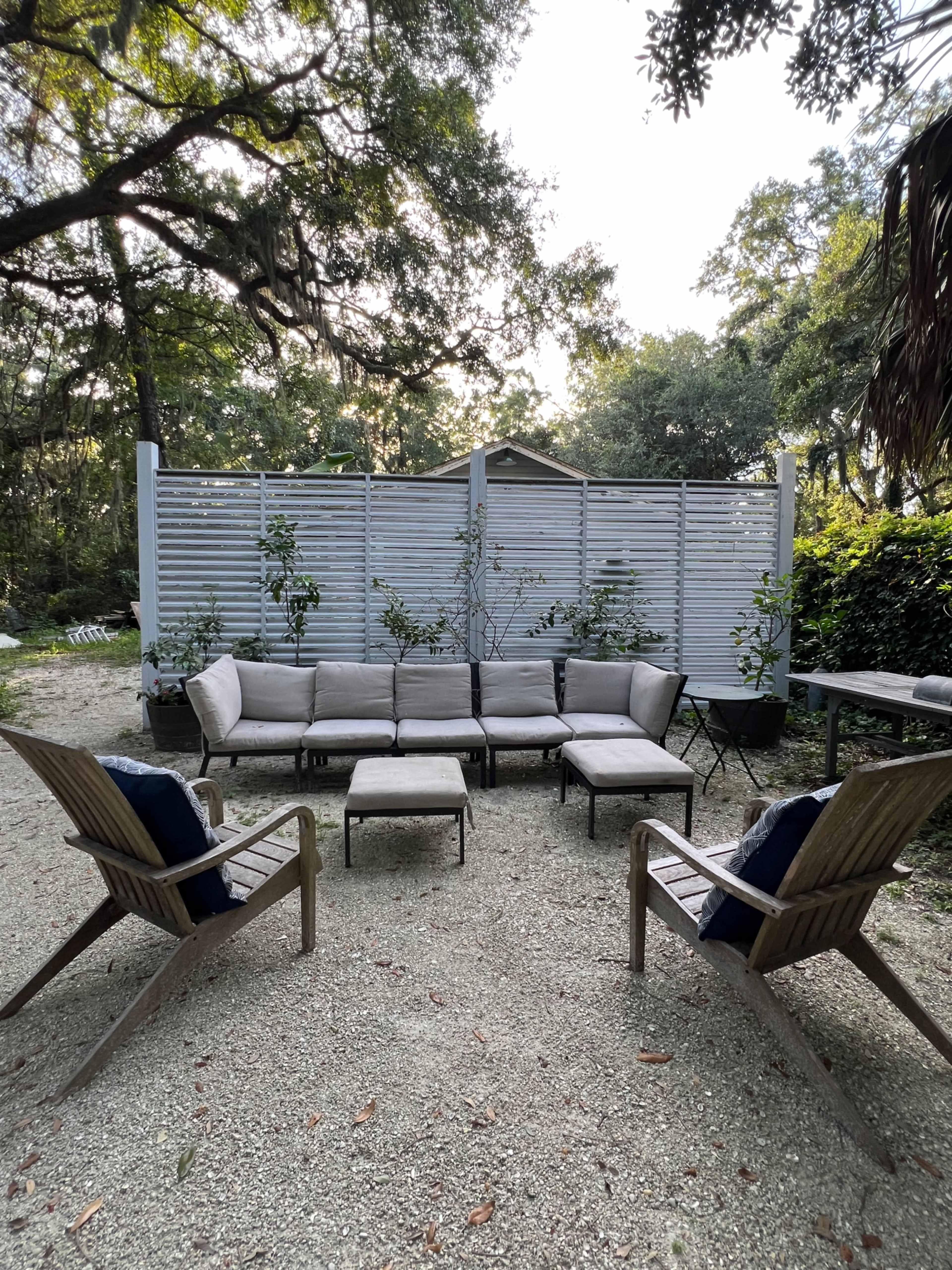 A cozy outdoor seating area features a sectional couch, two armchairs, and a backdrop of greenery and a wooden lattice fence.