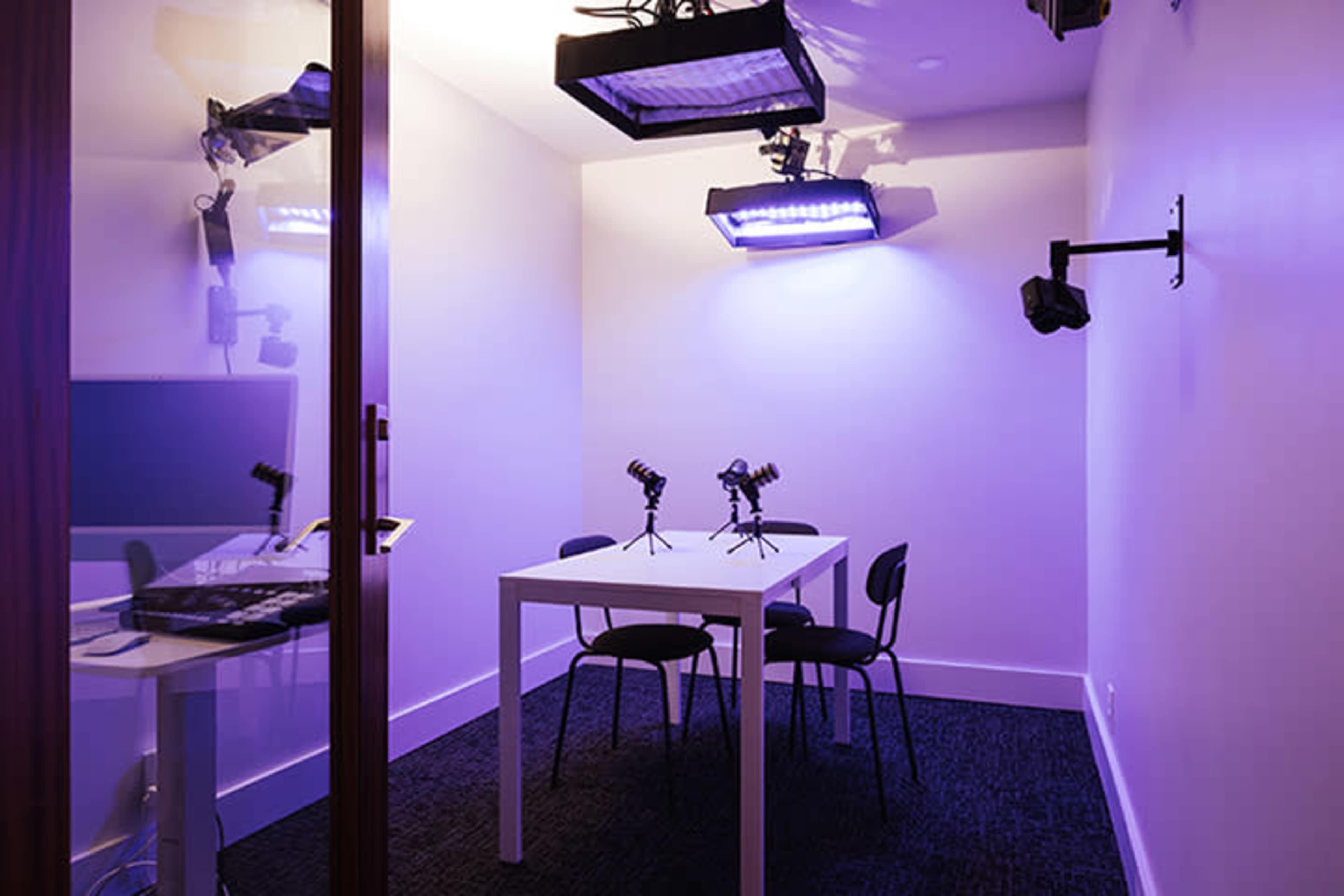The image shows a small, brightly lit room with a table and four chairs, equipped with several mounted lights and microphones.