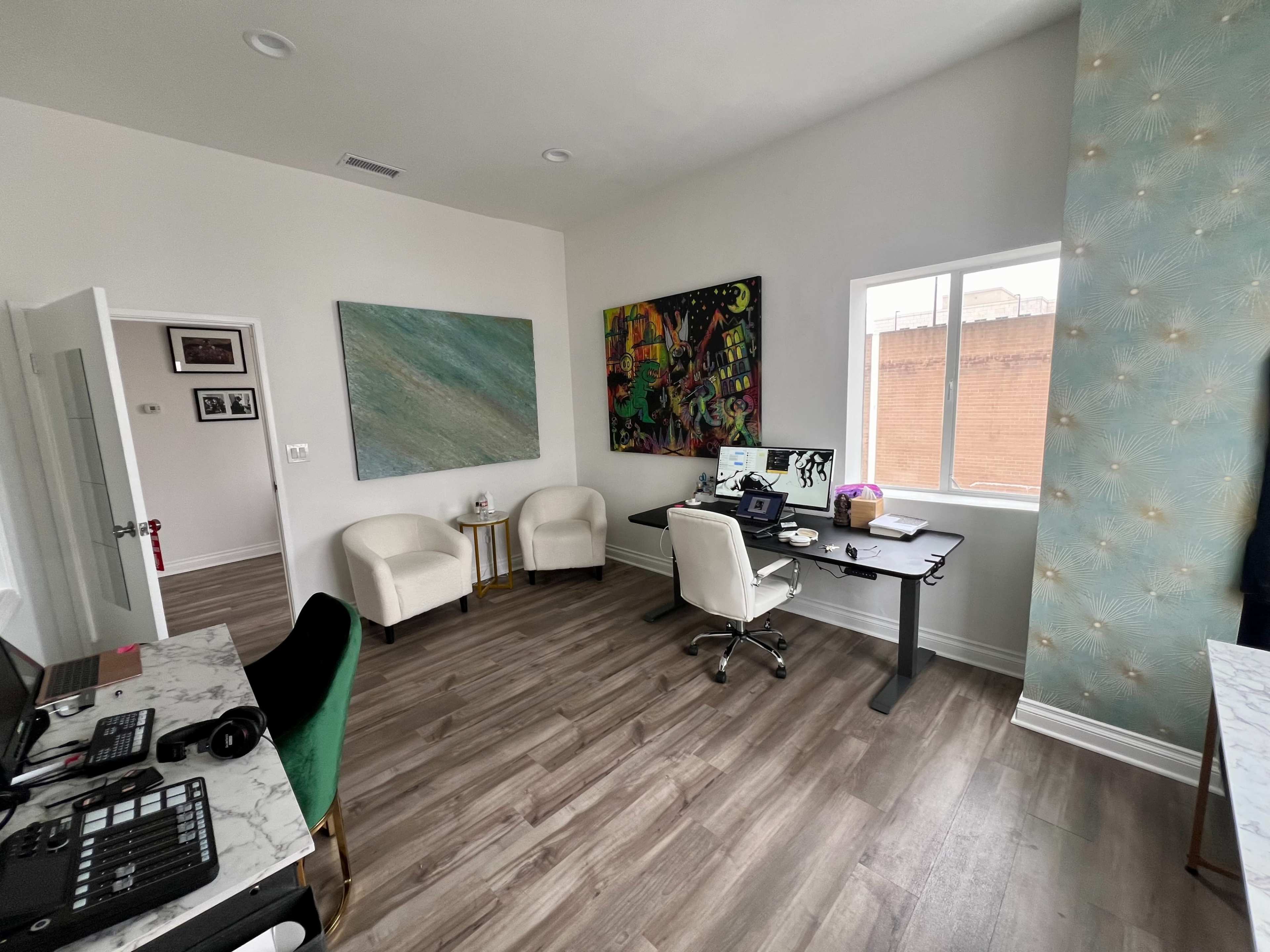 A modern office space featuring a desk with a computer, two white chairs, and colorful artwork on the walls.