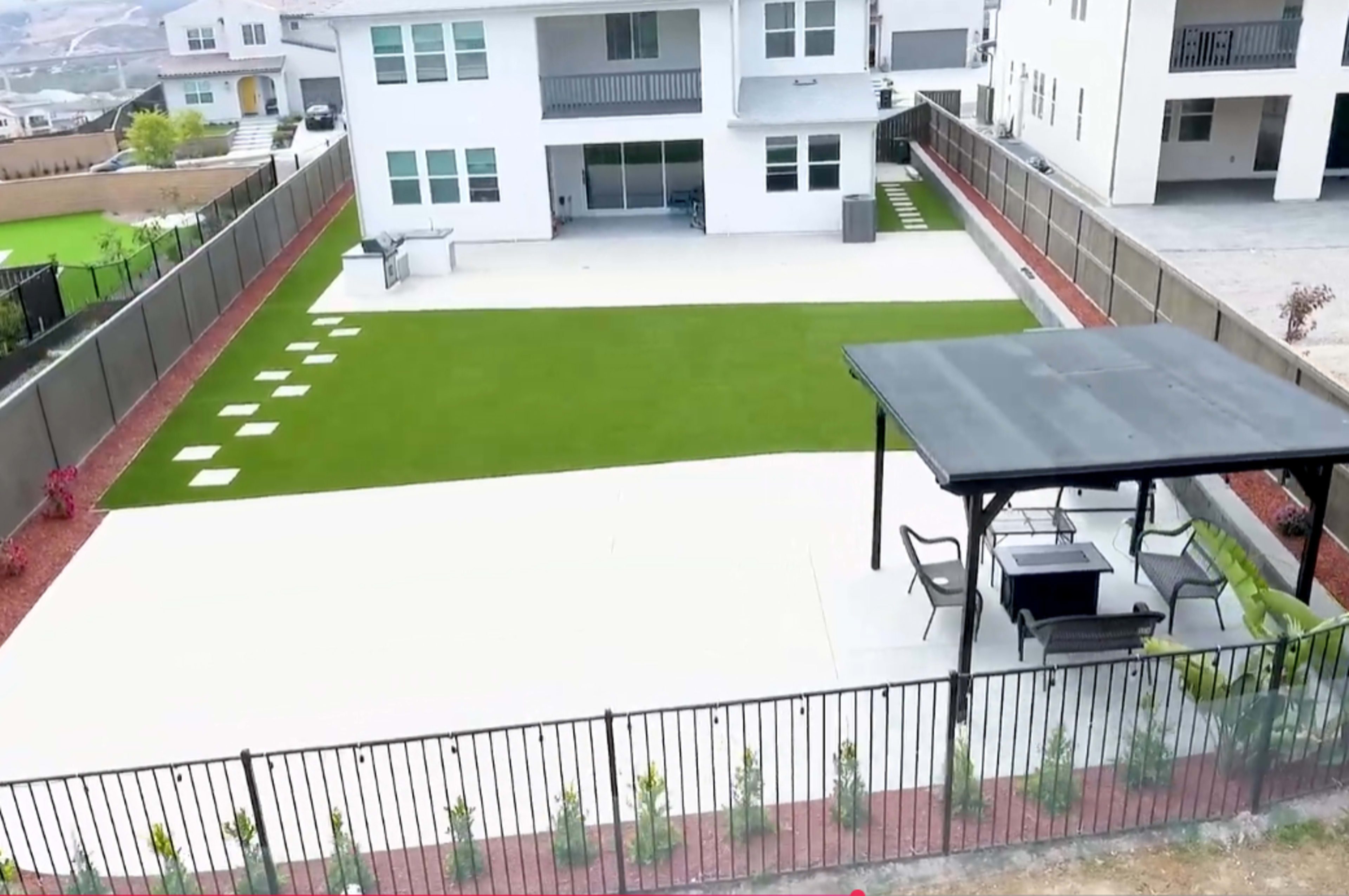 The image shows a large, landscaped backyard with a green artificial lawn, paved areas, and a shaded seating structure, surrounded by a black fence.