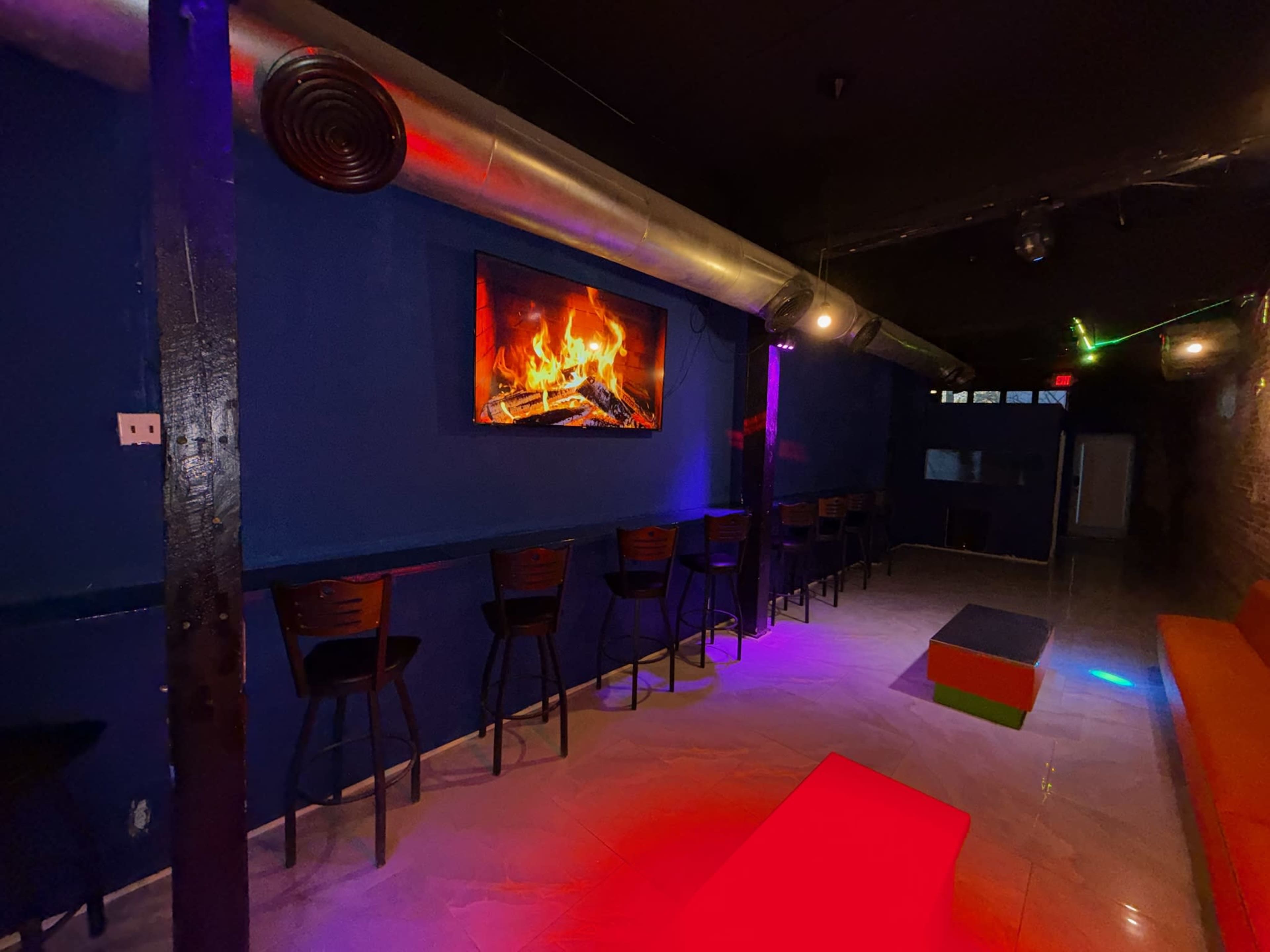The image shows a dimly lit bar area featuring a long row of black stools against a blue wall, with a fireplace-themed screen on display and colorful lighting effects.