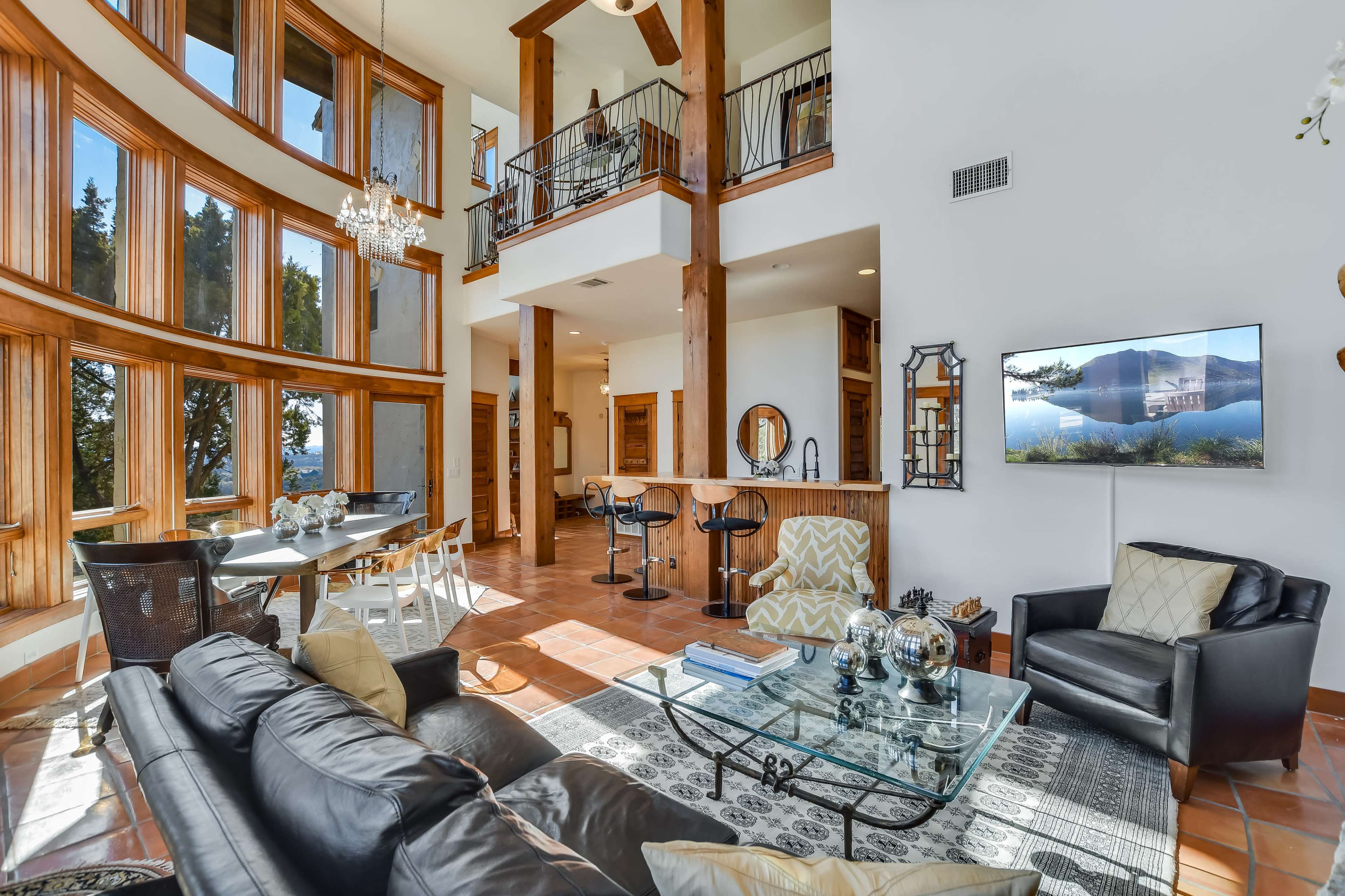 The image shows a spacious living area featuring large windows, wooden beams, a dining table, and a leather sofa with a television mounted on the wall.