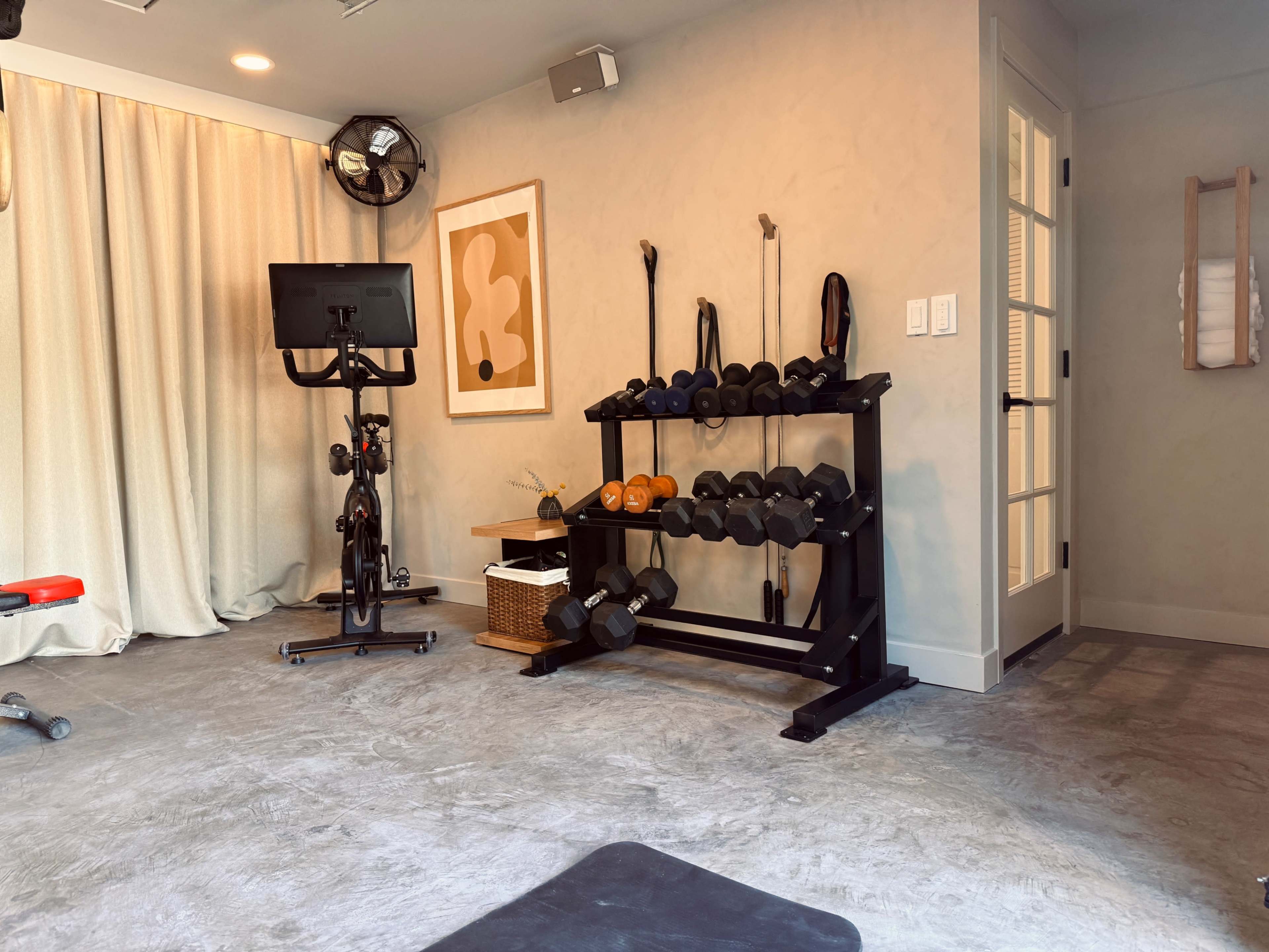 The image shows a home gym with a weight rack filled with dumbbells, a stationary bike, and a wall adorned with a framed artwork and various exercise accessories.