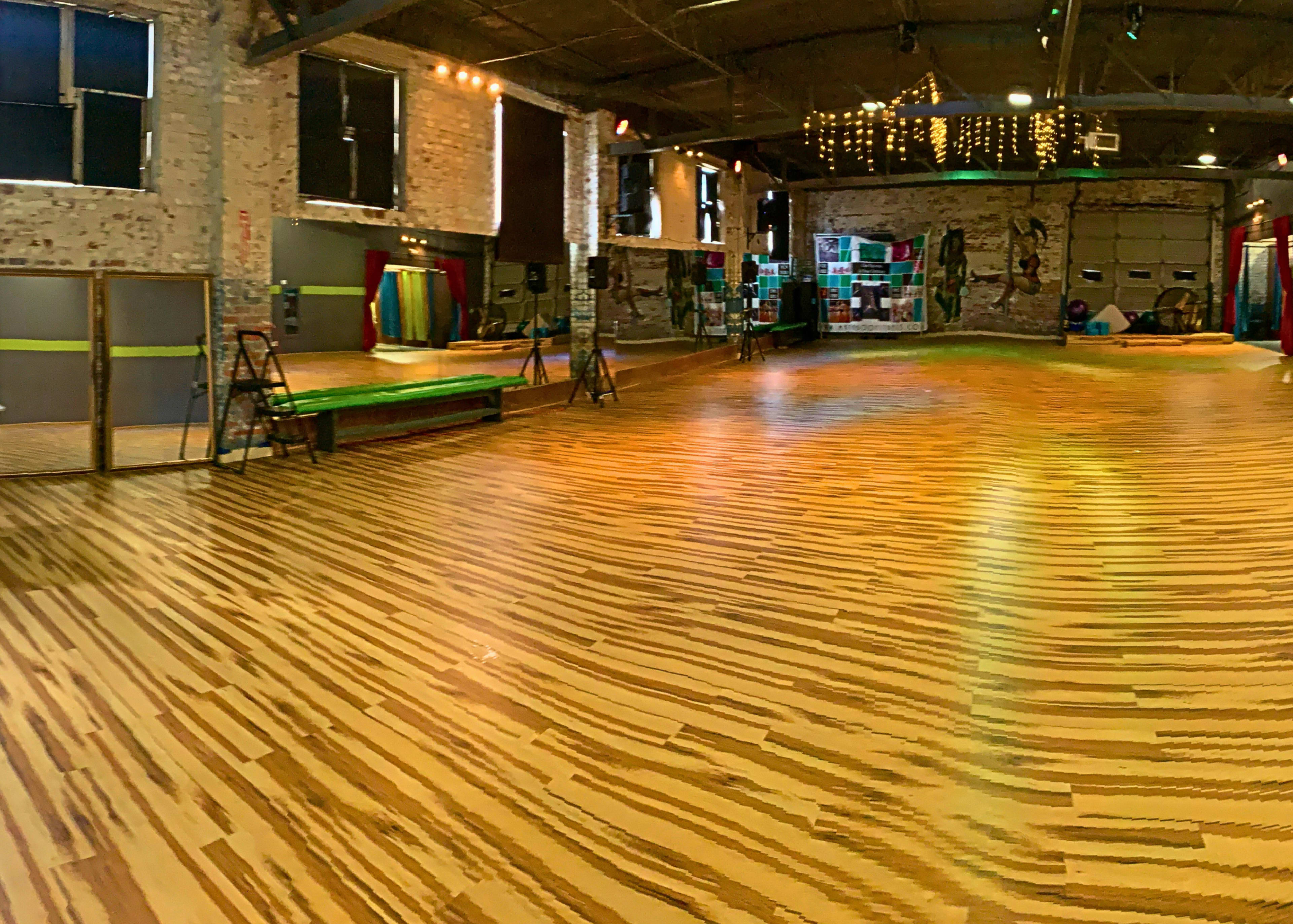 A spacious wooden-floored dance studio with exposed brick walls and various props along the sides.
