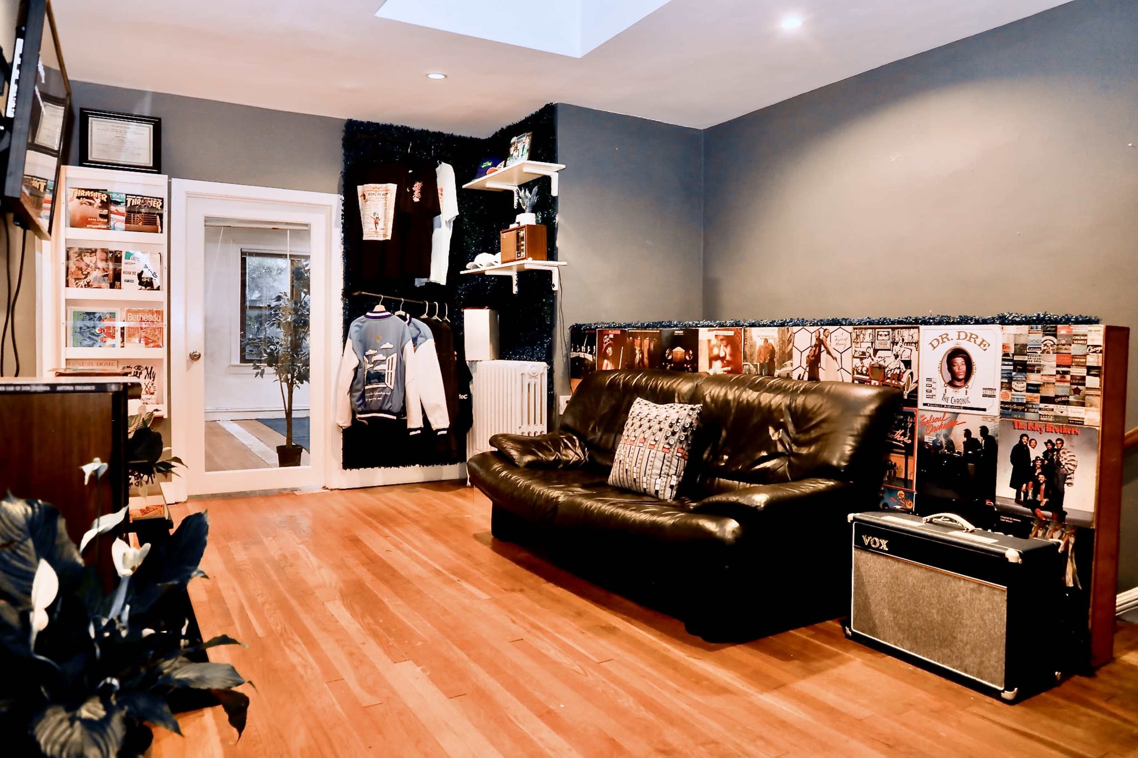 The image shows a cozy room with wooden floors, featuring a black leather sofa, a music amplifier, and walls decorated with framed posters and shelves.