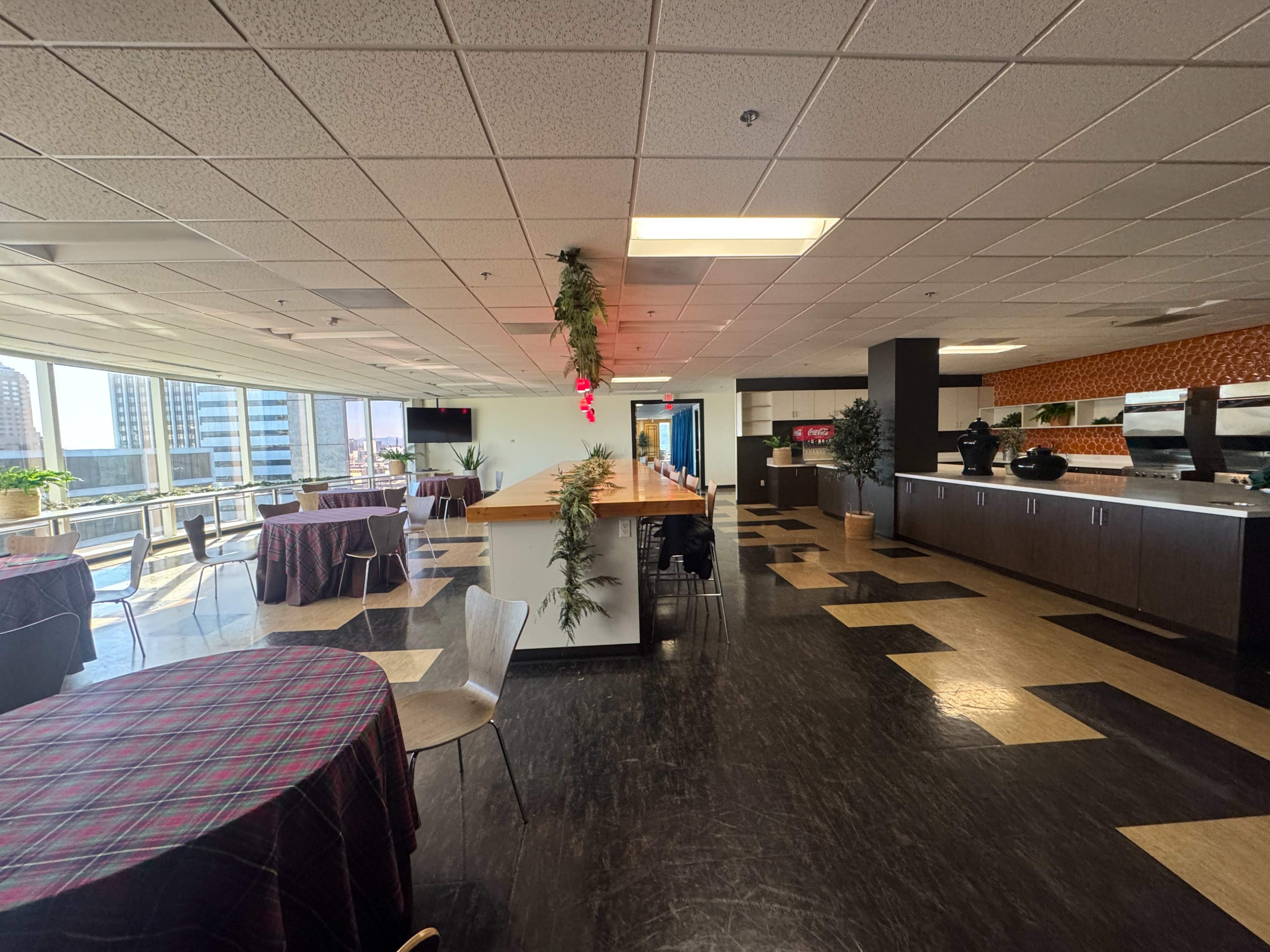 The image shows a spacious event space with tables set up and a modern kitchen area in the background, featuring large windows that provide a view of the city.