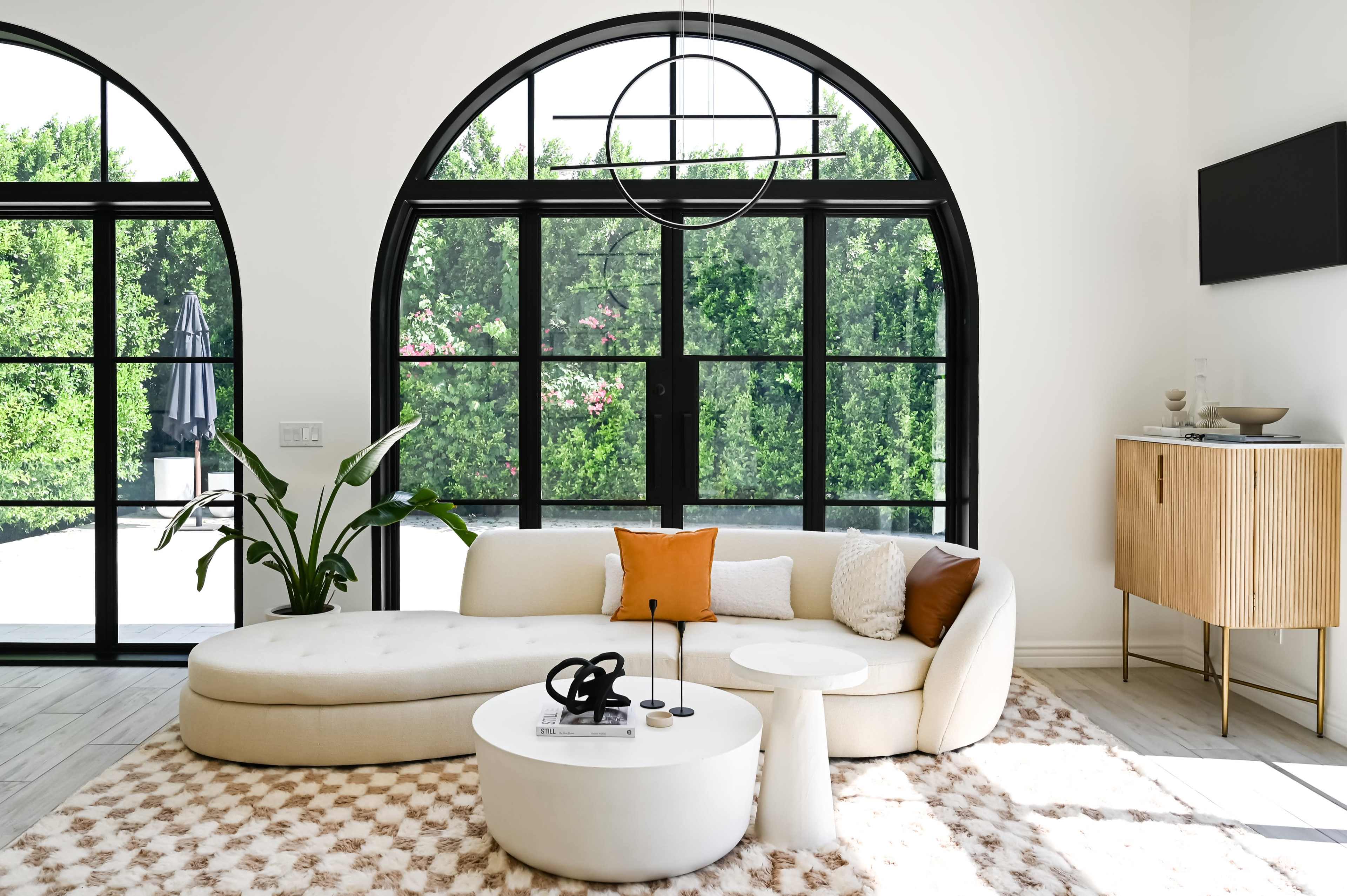 A modern living room featuring a curved sofa, a circular coffee table, and large arched windows that provide views of greenery outside.