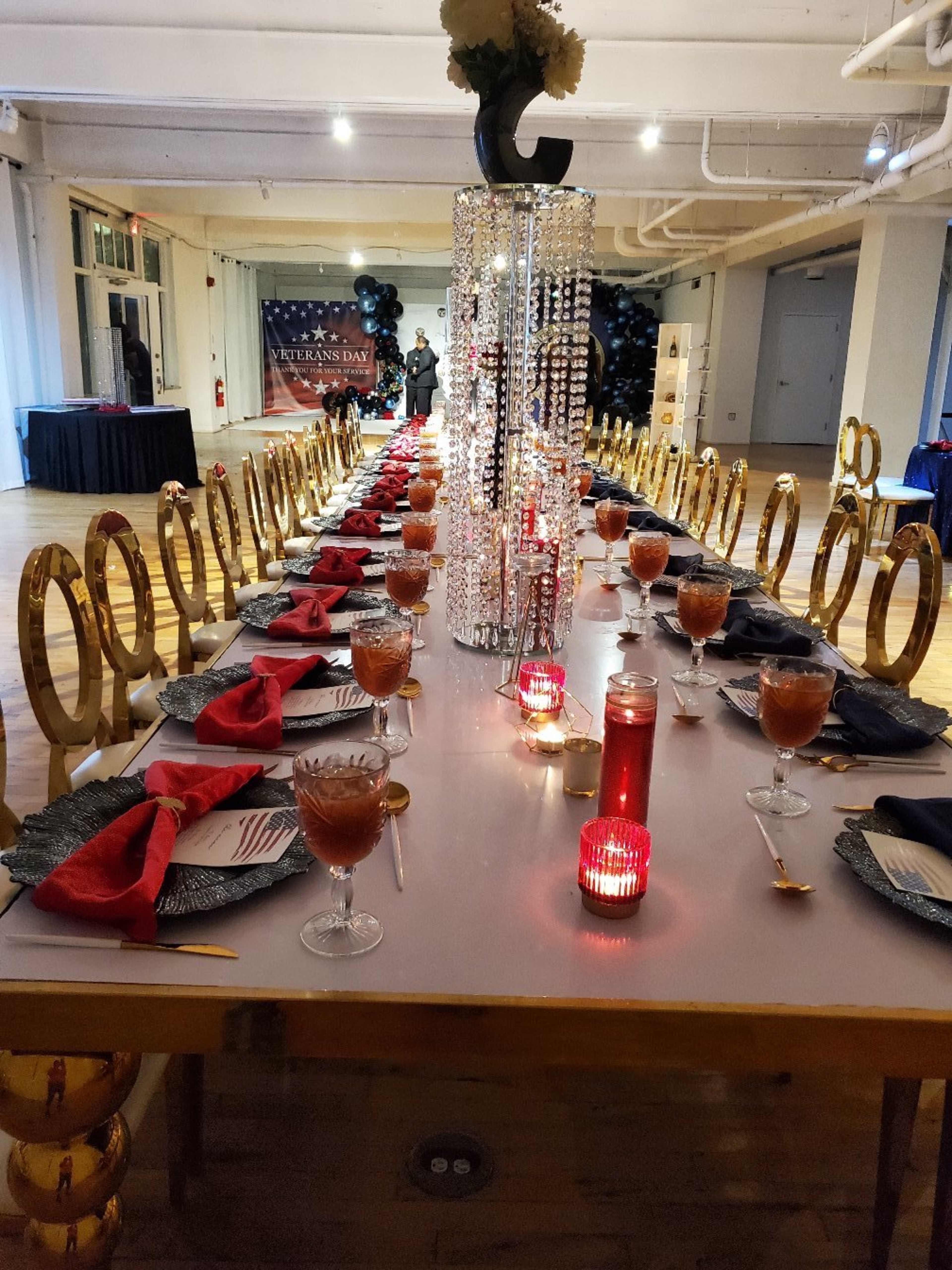 A long banquet table is set for an event, adorned with red napkins, candles, and decorative glassware, in a well-lit hall decorated with balloons and a backdrop.