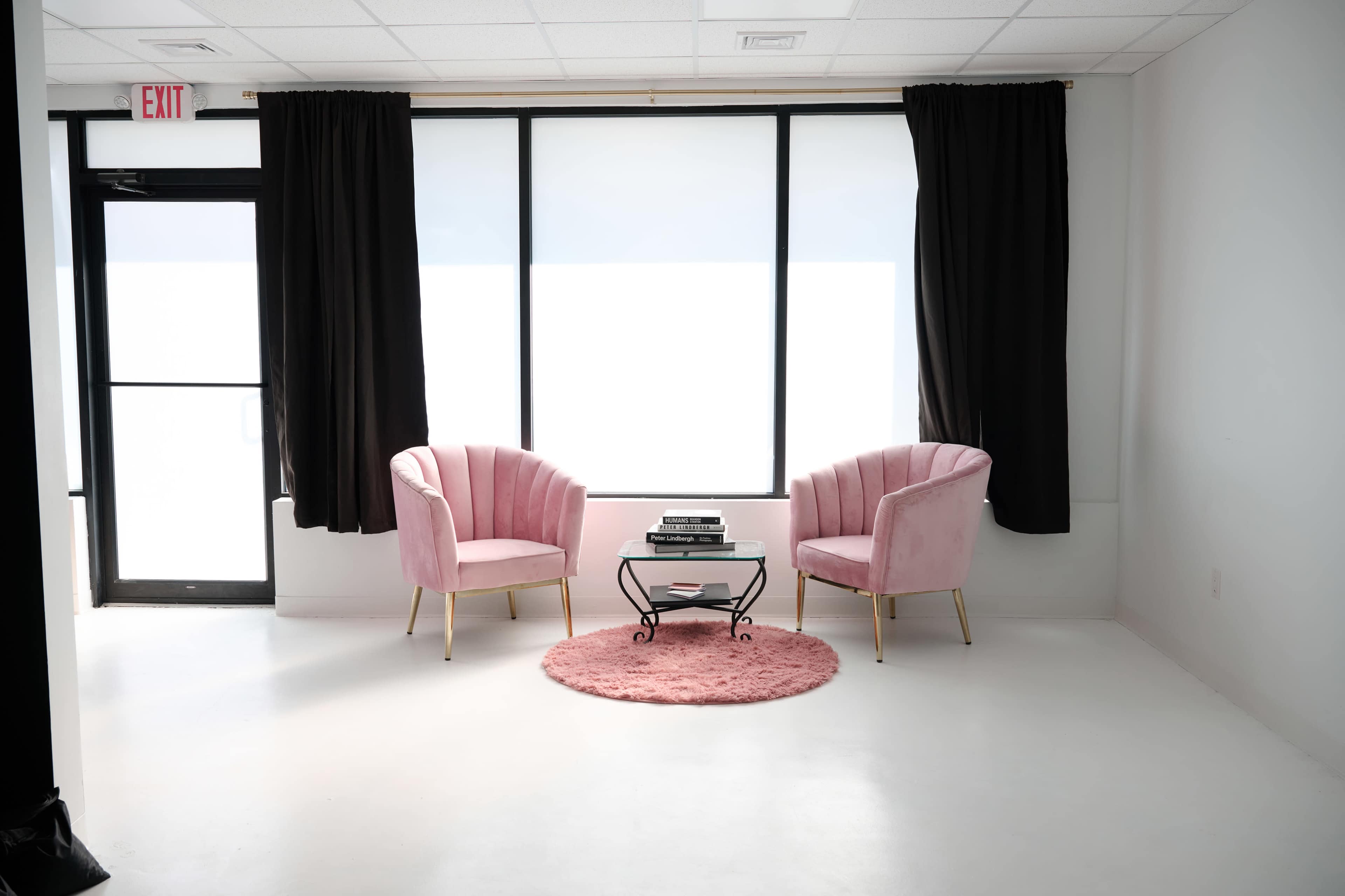 The image depicts a minimalist interior with two pink velvet chairs, a small black table, and a round pink rug, set near a large window draped with dark curtains.