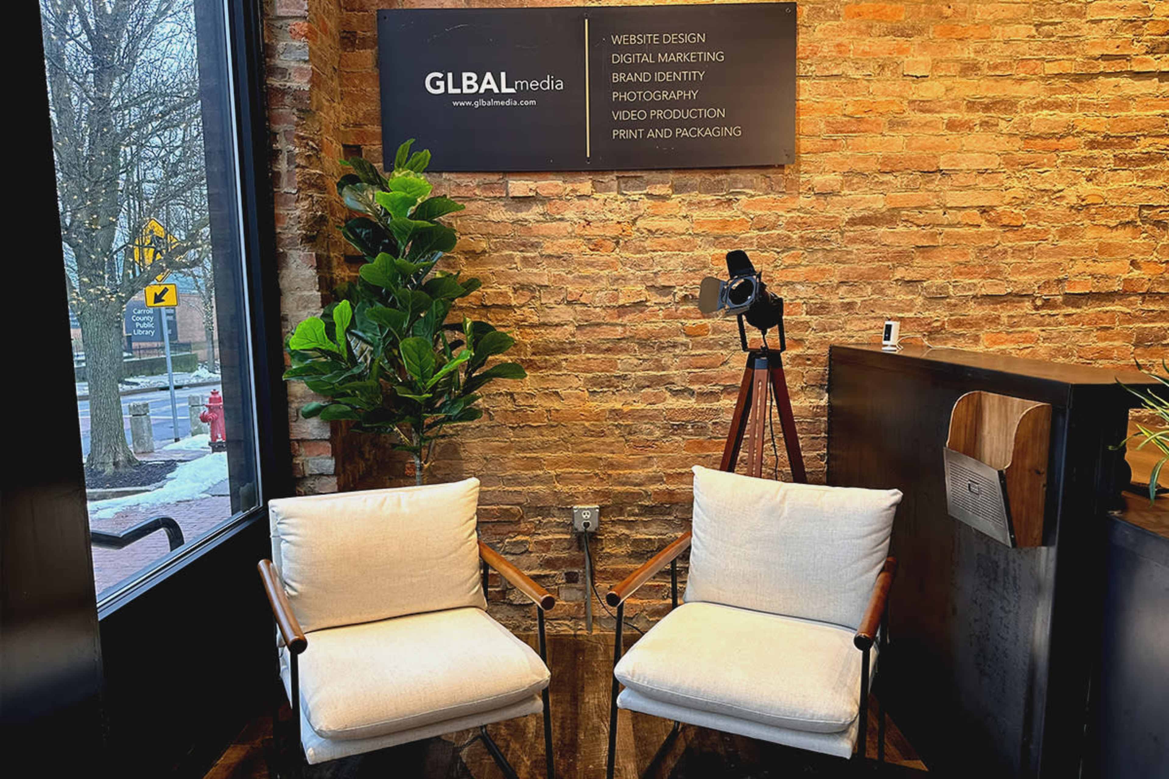 A modern workspace with two white chairs, a potted plant, and a wooden table under a sign displaying the services of a media company.
