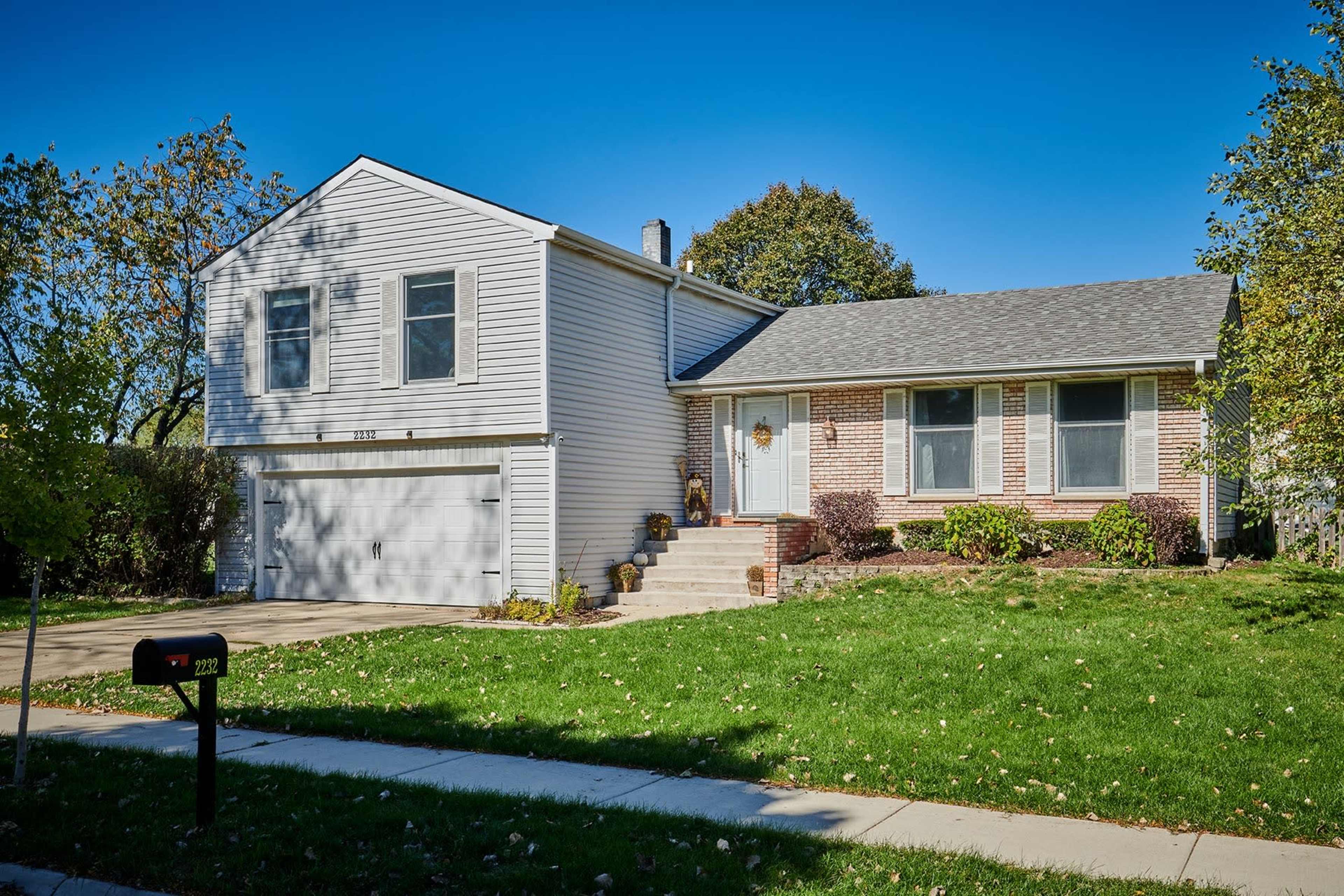 A two-story house with a brick front, gray roof, and a driveway leads up to a spacious lawn.