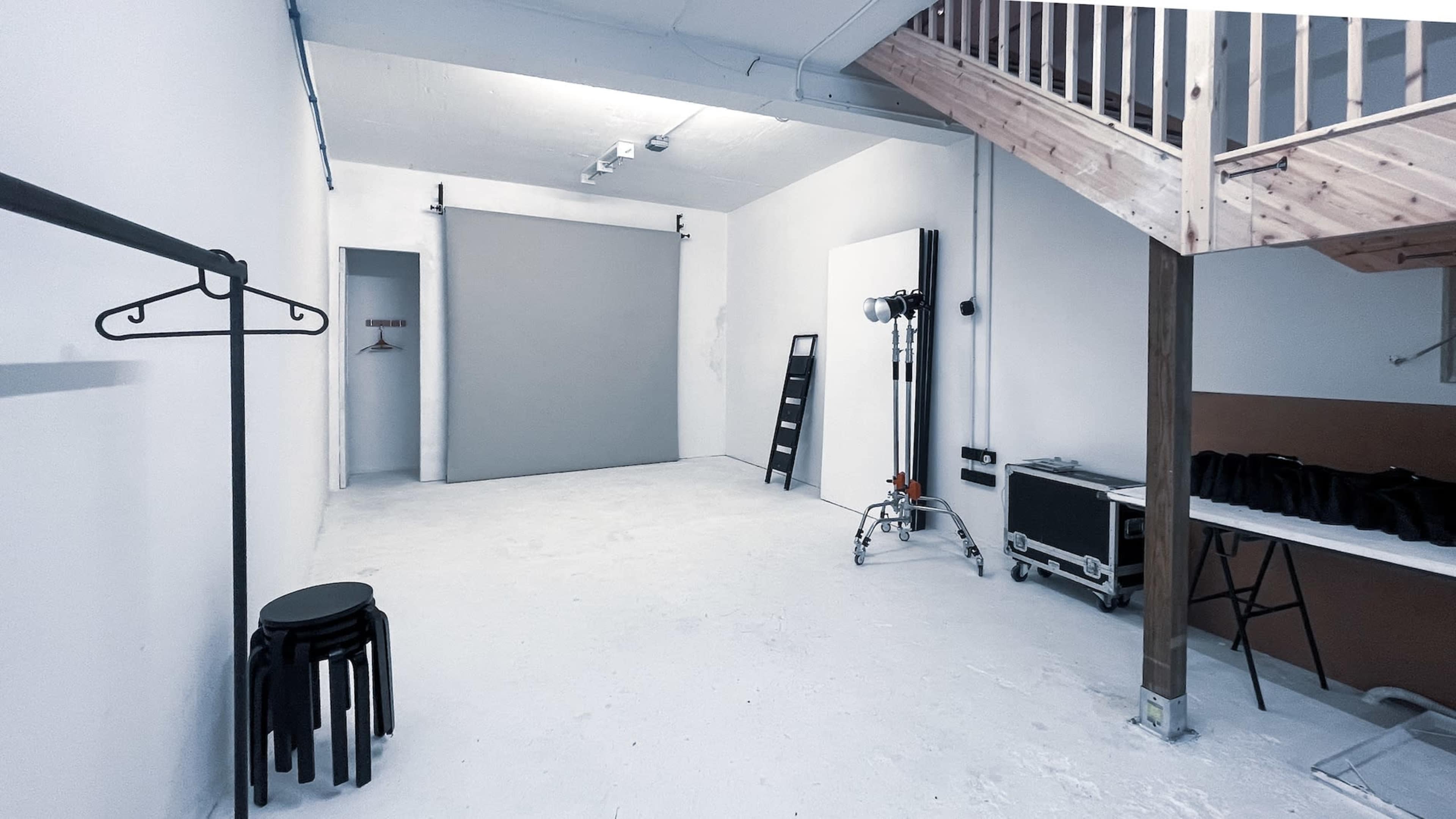 The image shows a spacious photography studio with a gray backdrop, a set of lights on a tripod, and various furniture pieces, including stools and a table along the side.