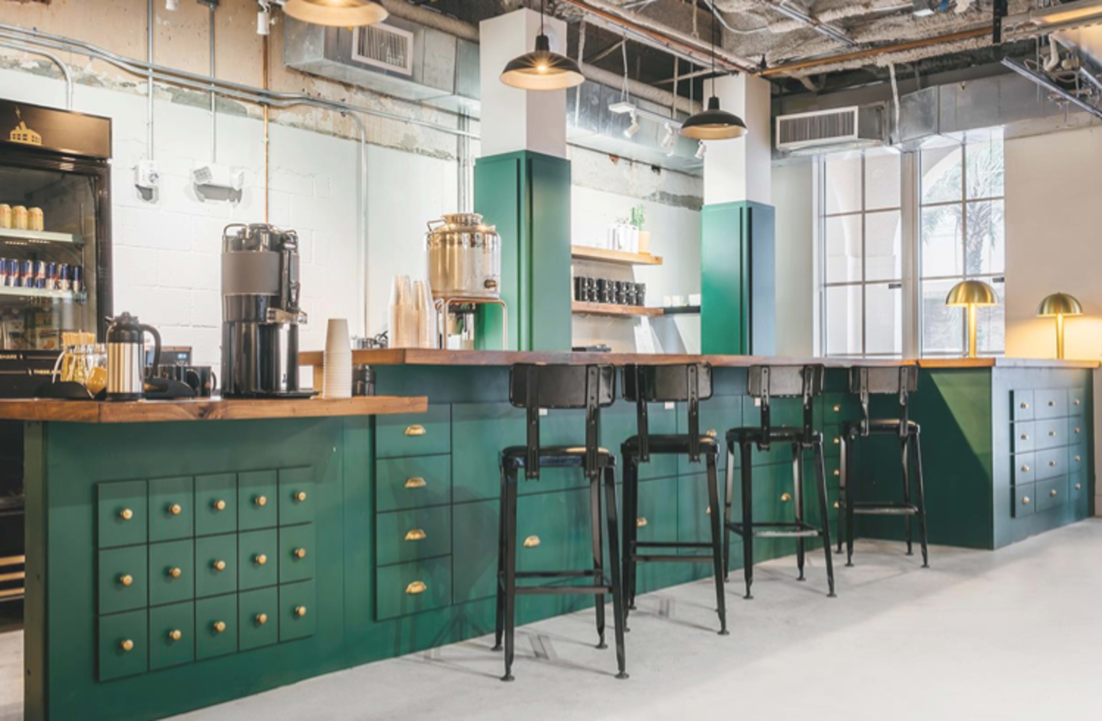 A modern coffee shop with a green and wooden bar, featuring high stools and decorative shelves.