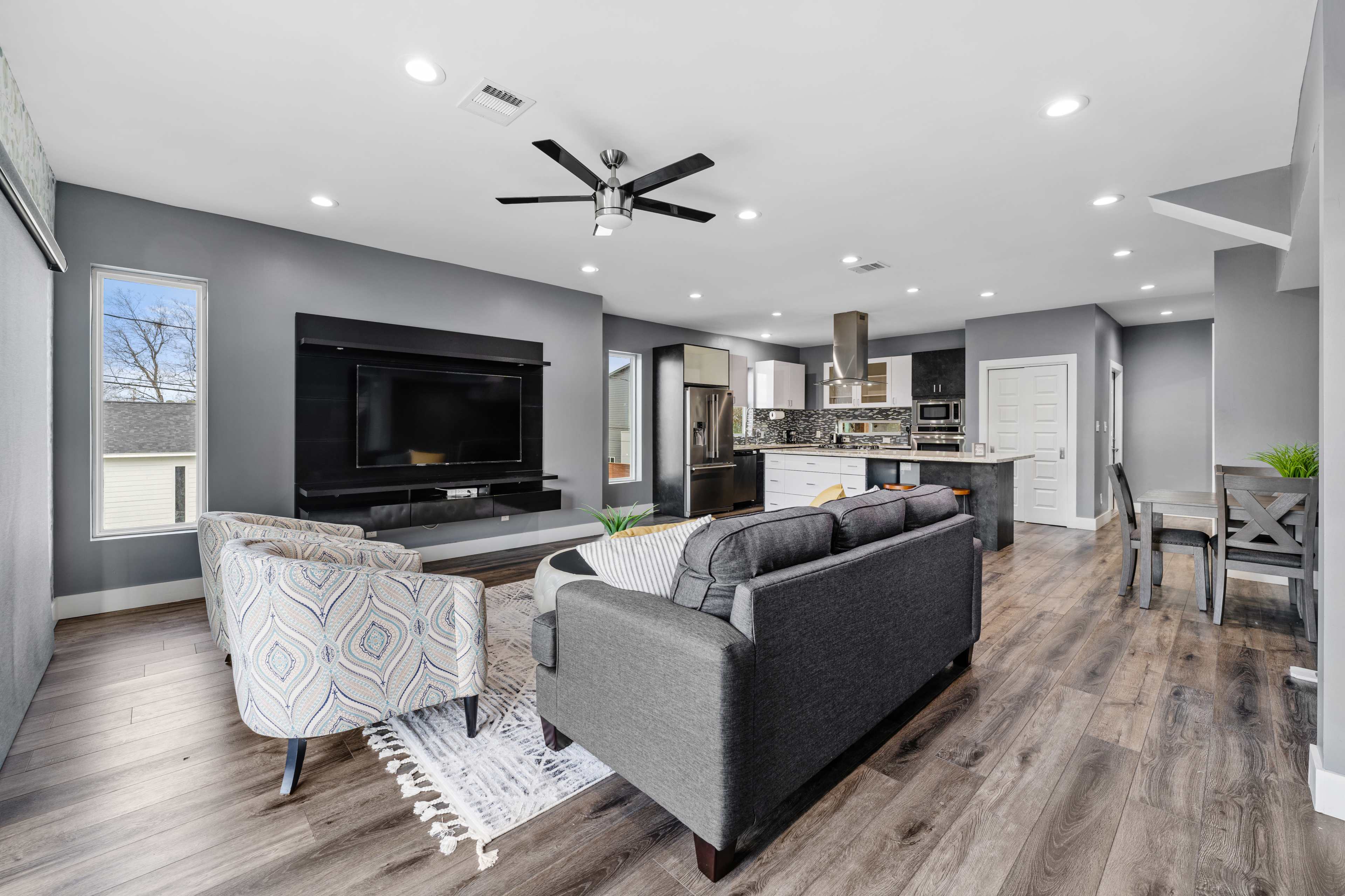 A modern living room features a dark TV wall unit, gray sofas, a patterned rug, and an open layout leading to a kitchen area.