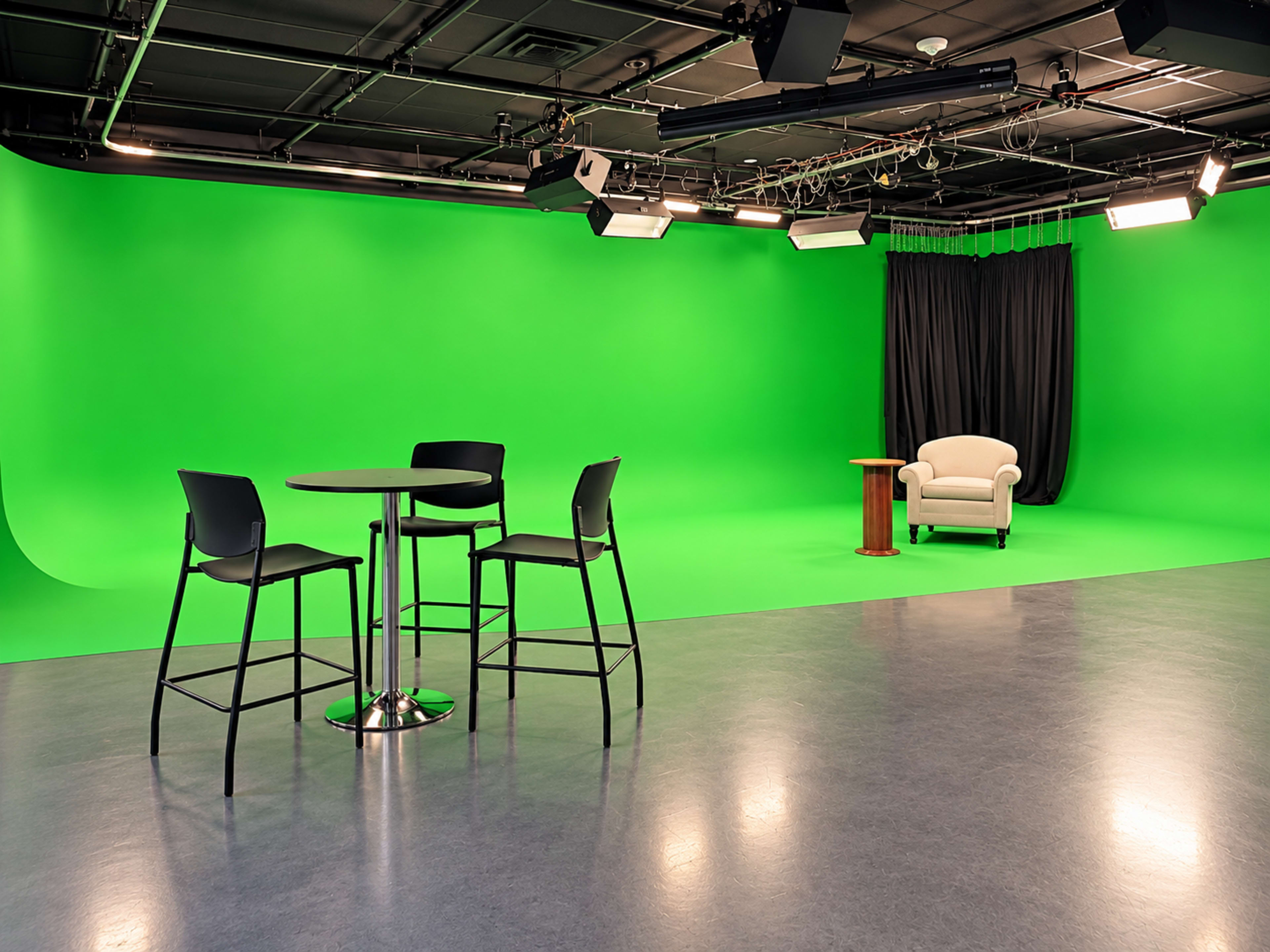 The image shows a studio space with a green screen backdrop, a circular table with four high chairs, and a small table next to a white armchair.