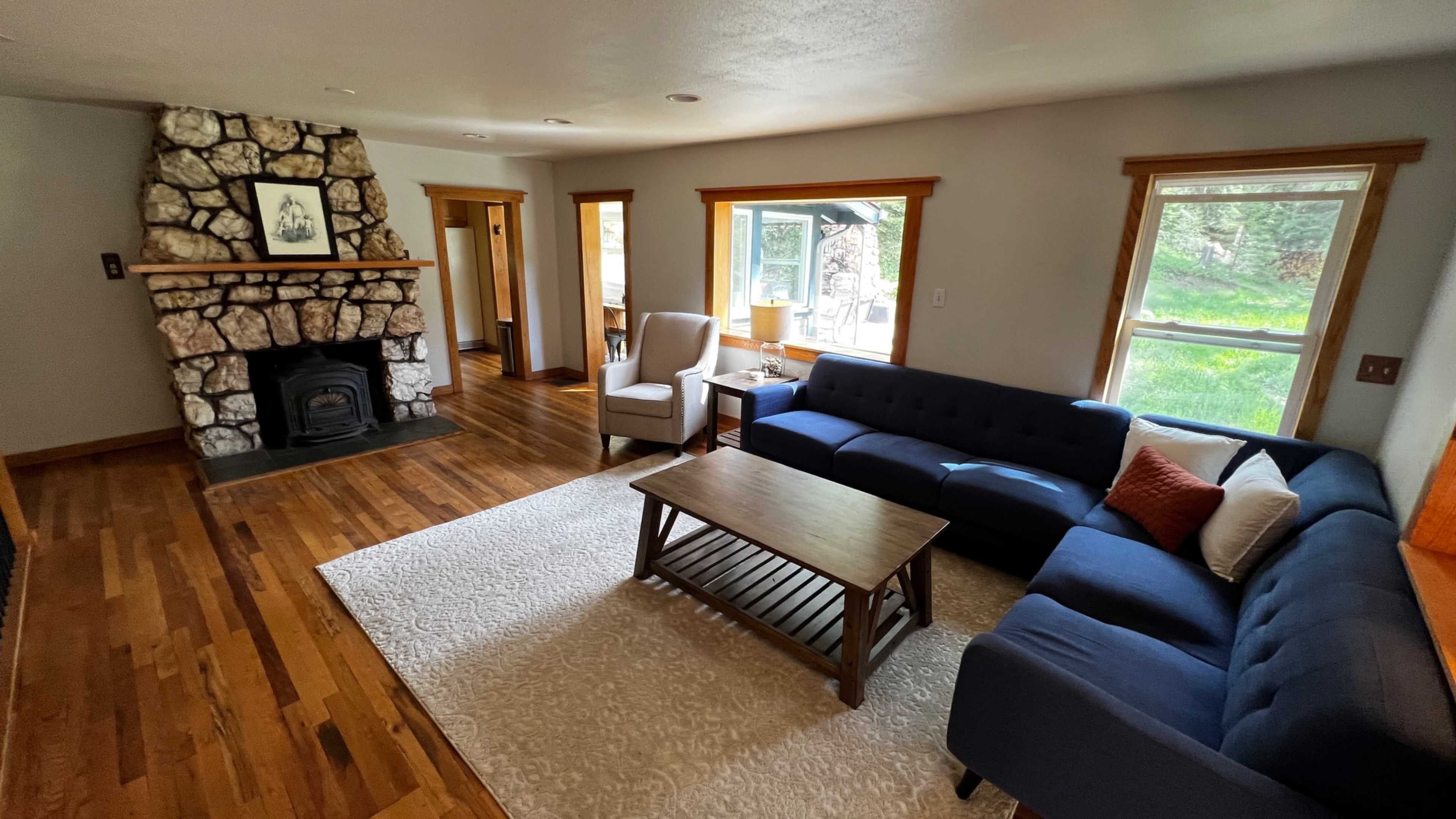 A living room with a stone fireplace, a blue sofa, a wooden coffee table, and large windows allowing natural light to enter.