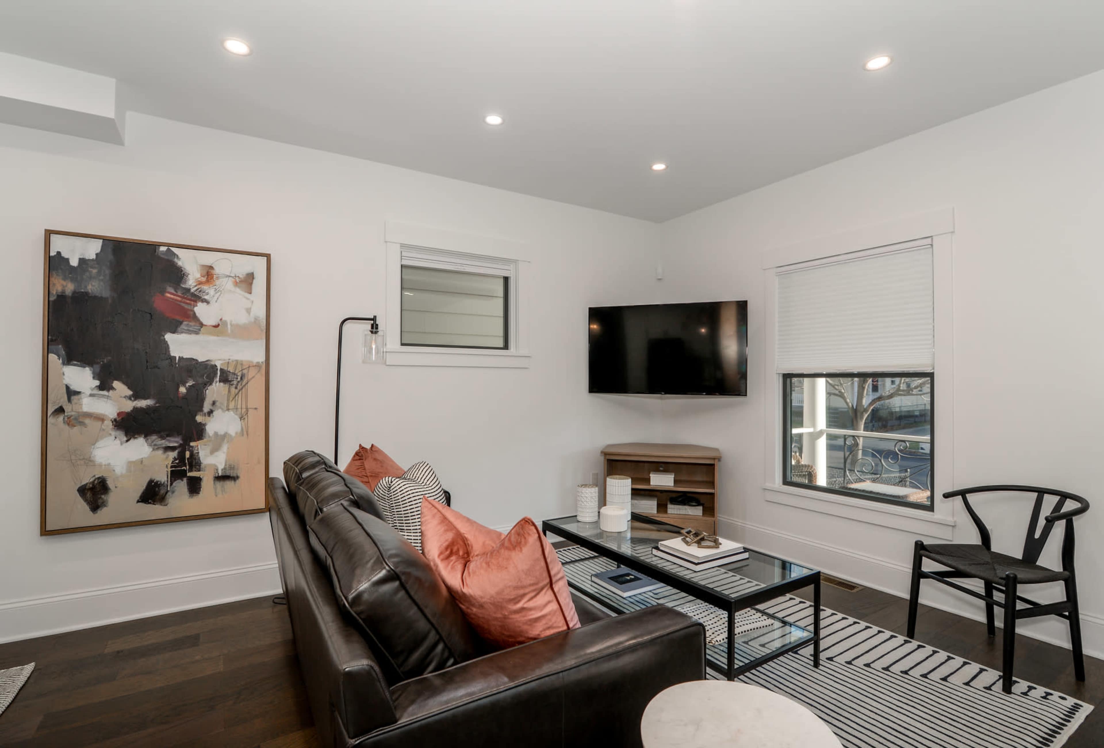 A modern living room features a dark leather sofa with orange pillows, a coffee table on a striped rug, a wall-mounted TV, and a large abstract painting.
