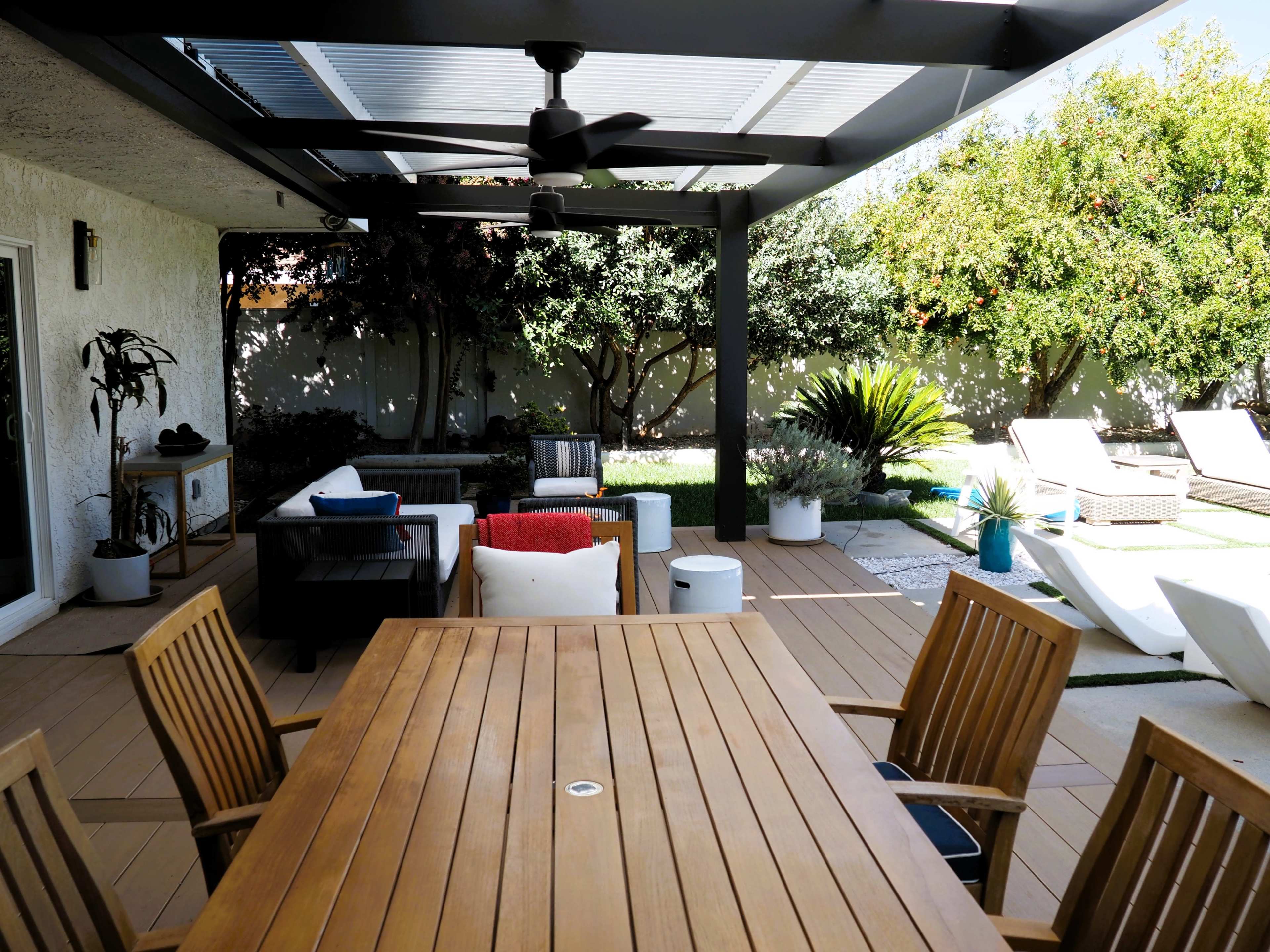 A wooden dining table with several chairs is positioned on a patio surrounded by plants and outdoor furniture beneath a covered pergola.