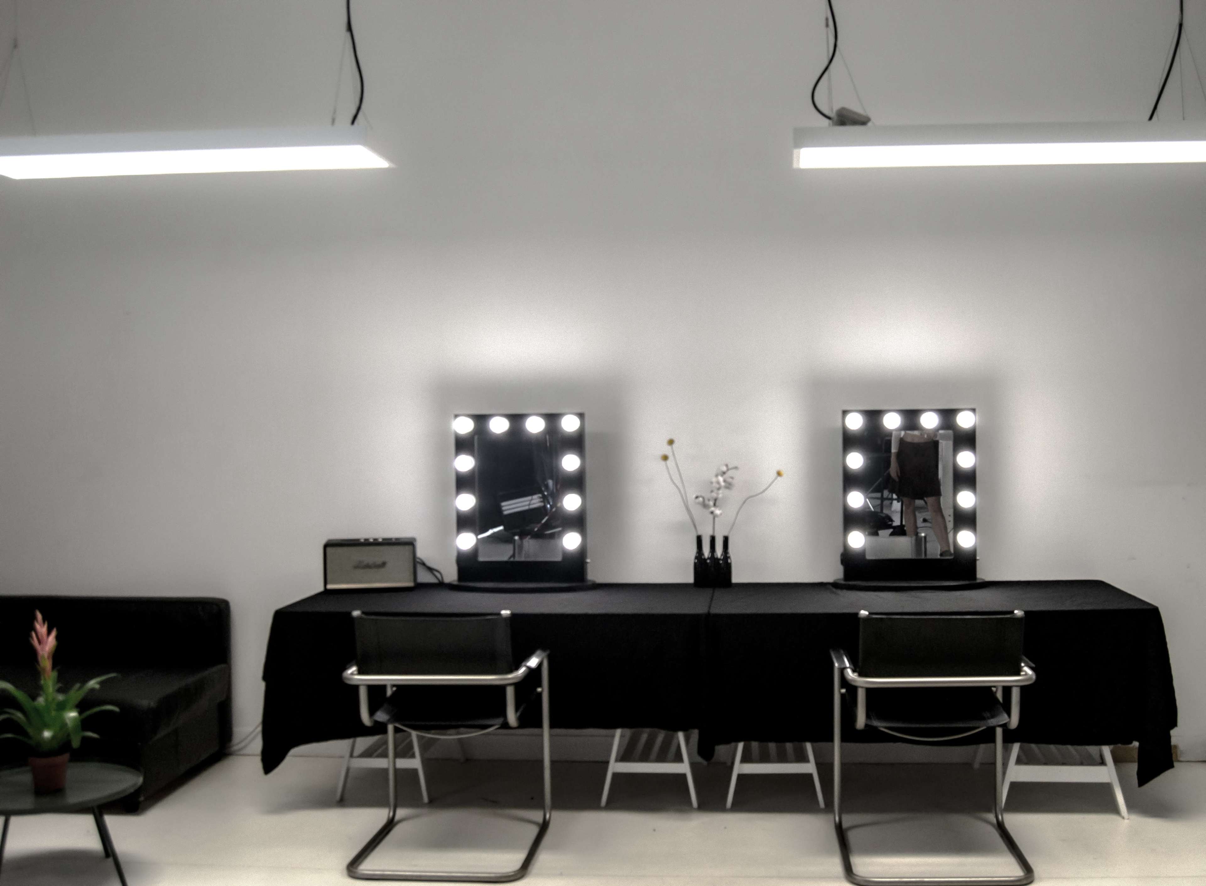 The image shows a minimalist salon space featuring two illuminated mirrors, a long black table, two chairs, and a decorative plant on one side.