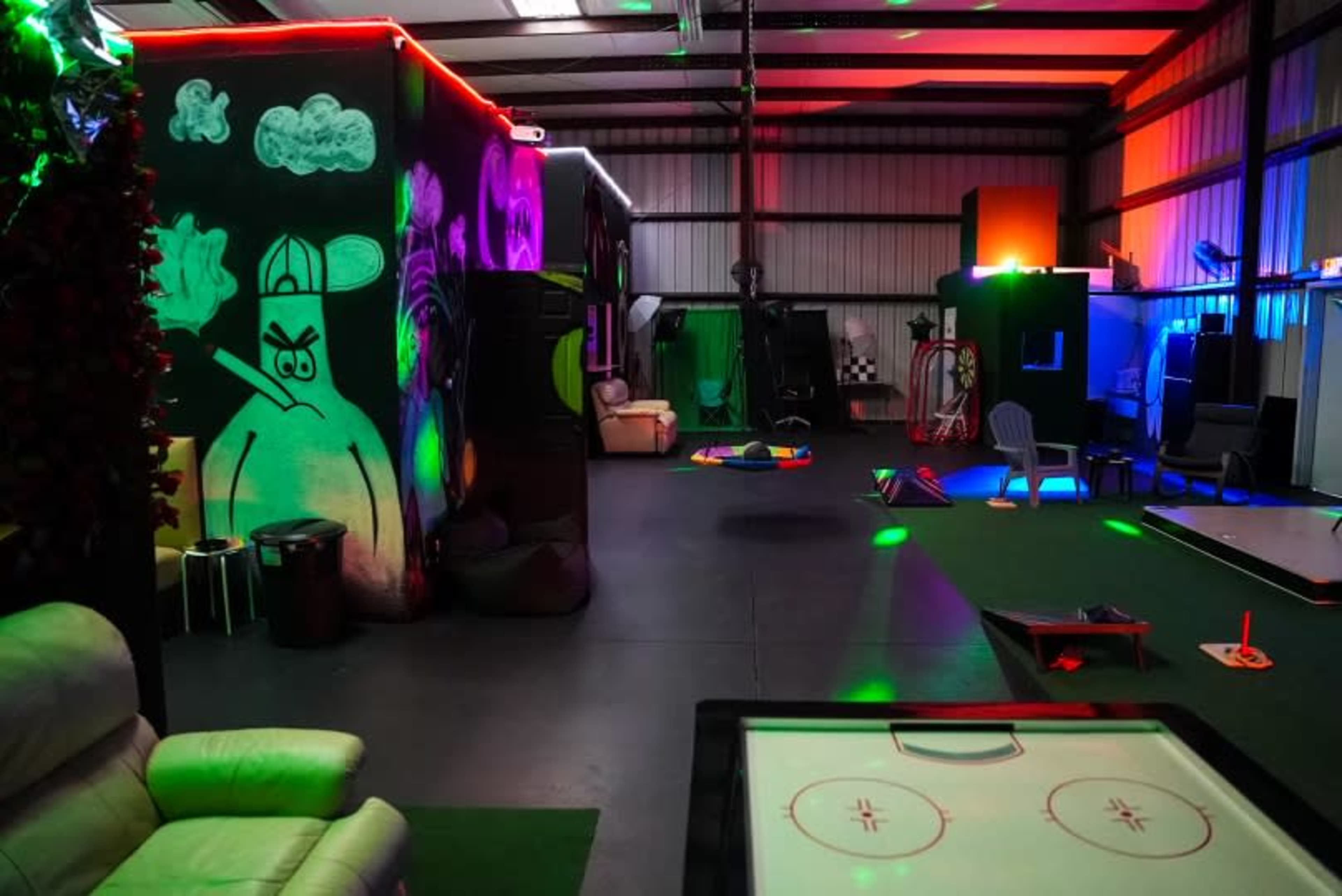 An indoor recreational space featuring colorful lighting, a mural on one wall, and various play areas with a foosball table and seating arrangements.