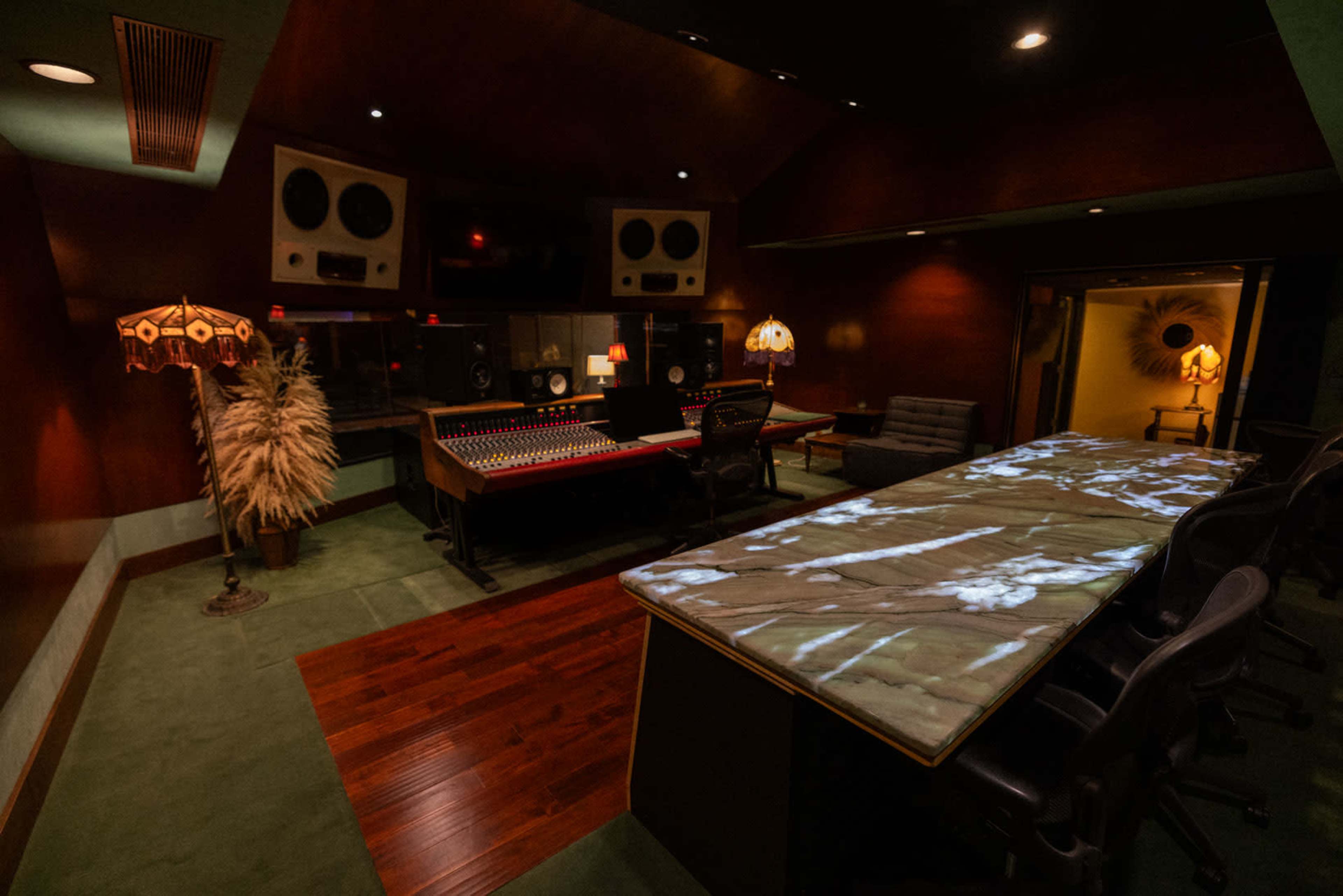 The image shows a dimly lit music studio featuring a large mixing console, a long illuminated table, and two decorative lamps in a room with wooden accents.