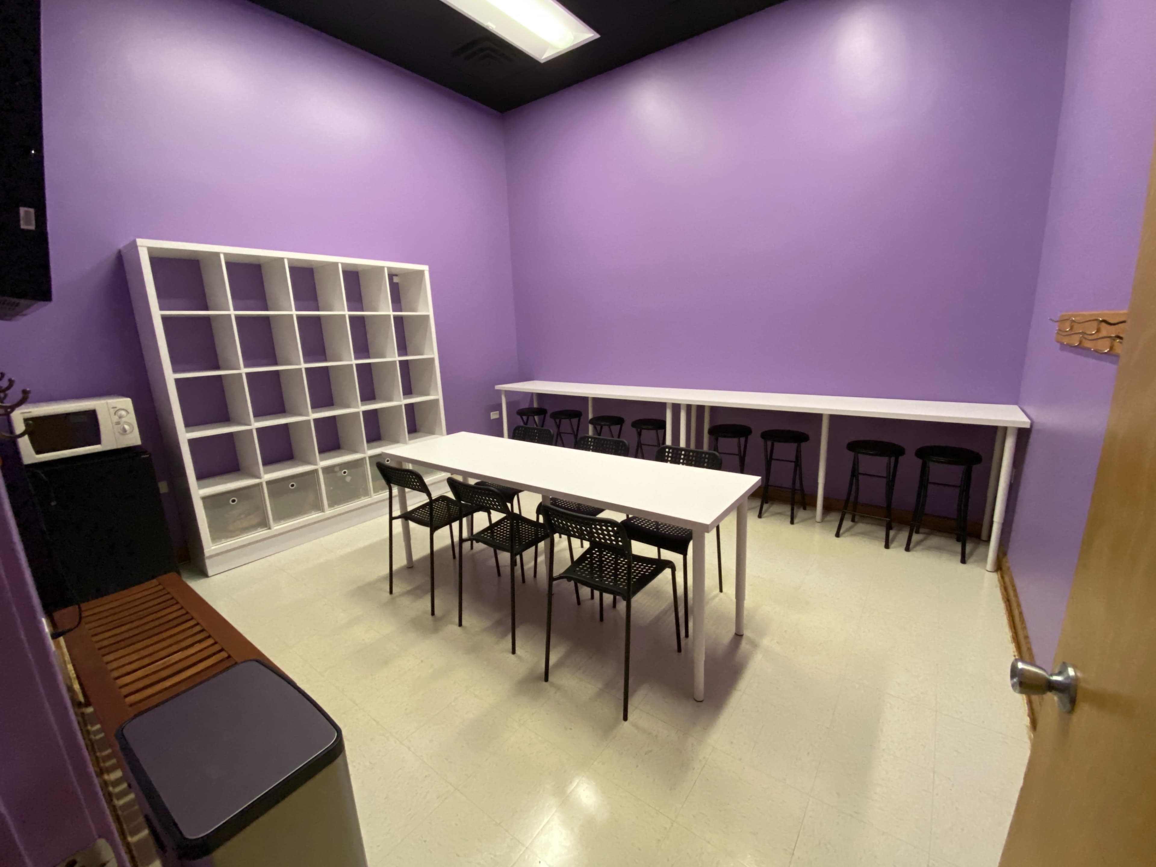 A room with purple walls, featuring a long white table surrounded by black chairs, a storage unit, and a kitchenette area.