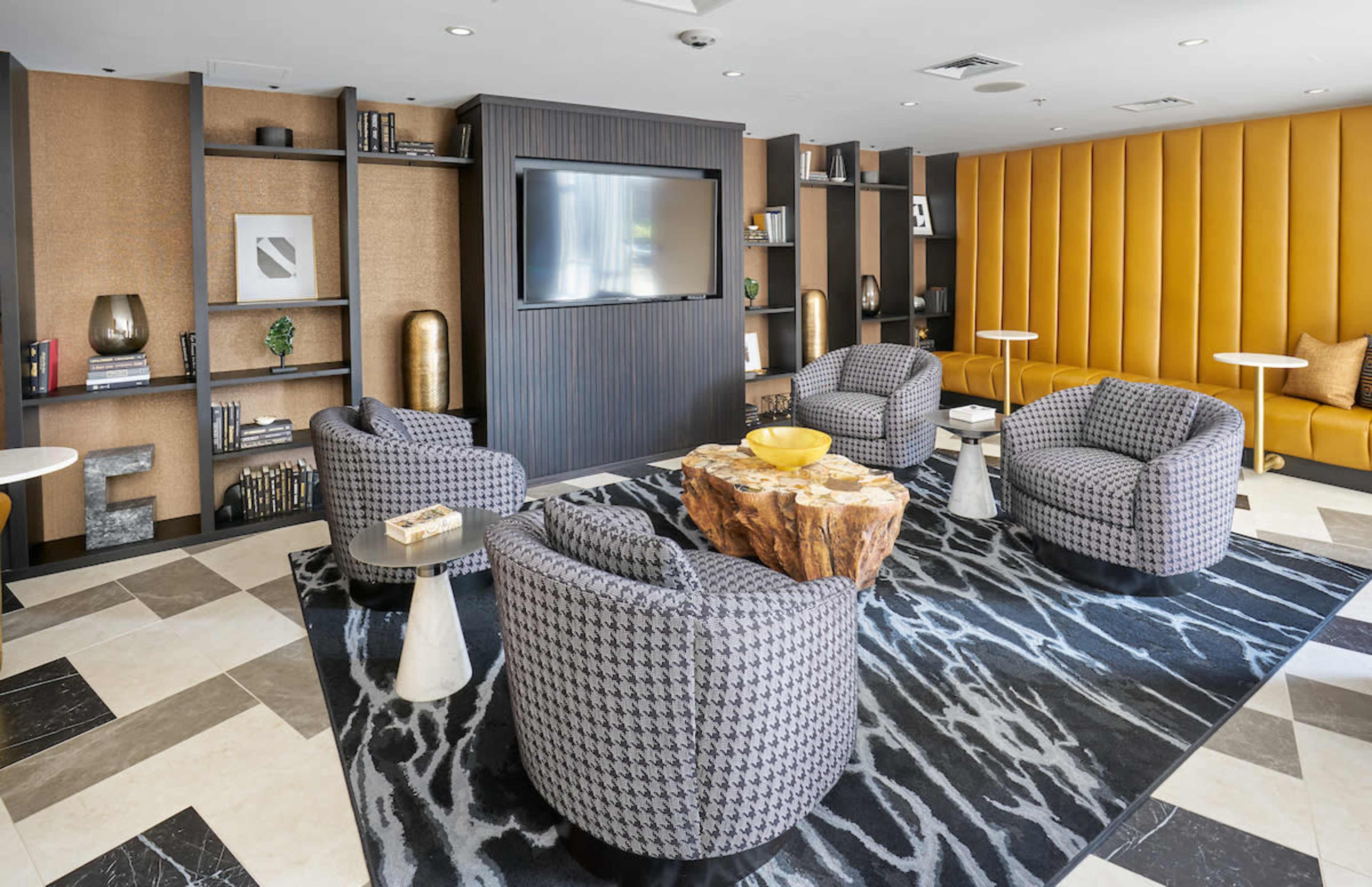 The image shows a modern lounge area featuring four patterned armchairs arranged around a wooden coffee table on a decorative rug, with a large flat-screen TV mounted on the wall and bookshelves in the background.