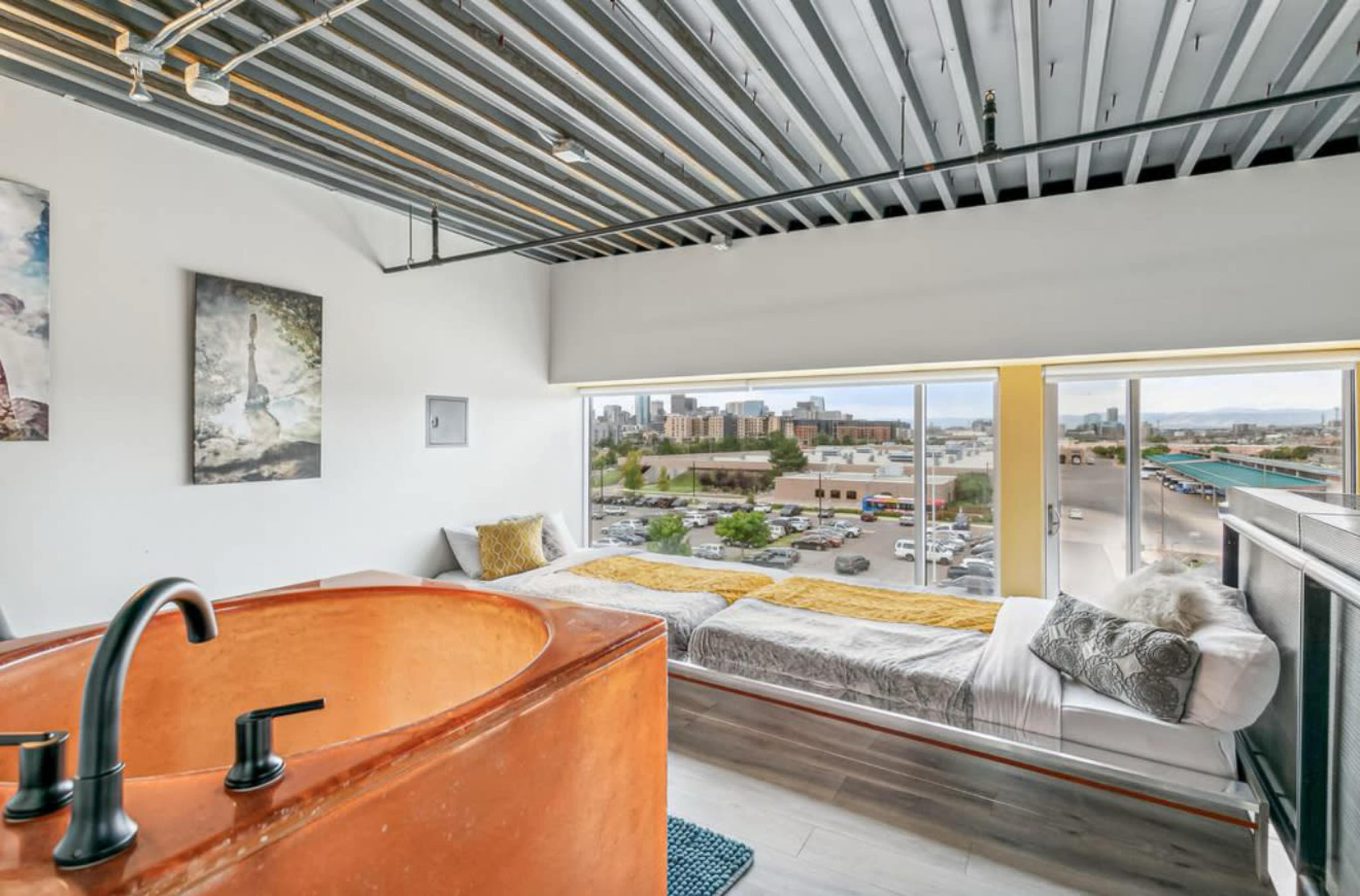 The image shows a modern bedroom with large windows overlooking a cityscape, featuring a unique orange sink and two beds adorned with yellow linens.