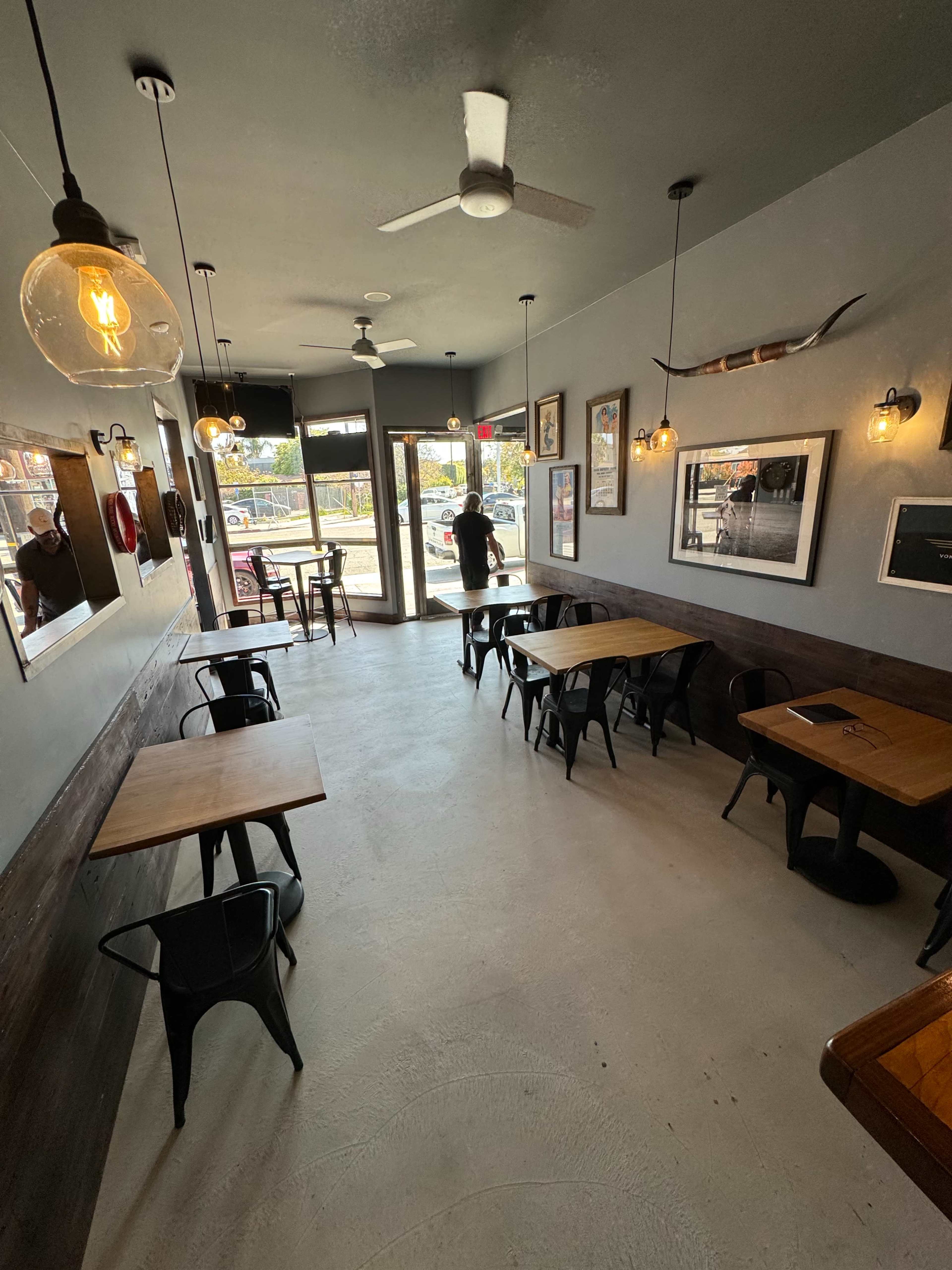 The image shows a well-lit café interior with wooden tables and metal chairs, featuring large windows that provide a view of the outdoors.