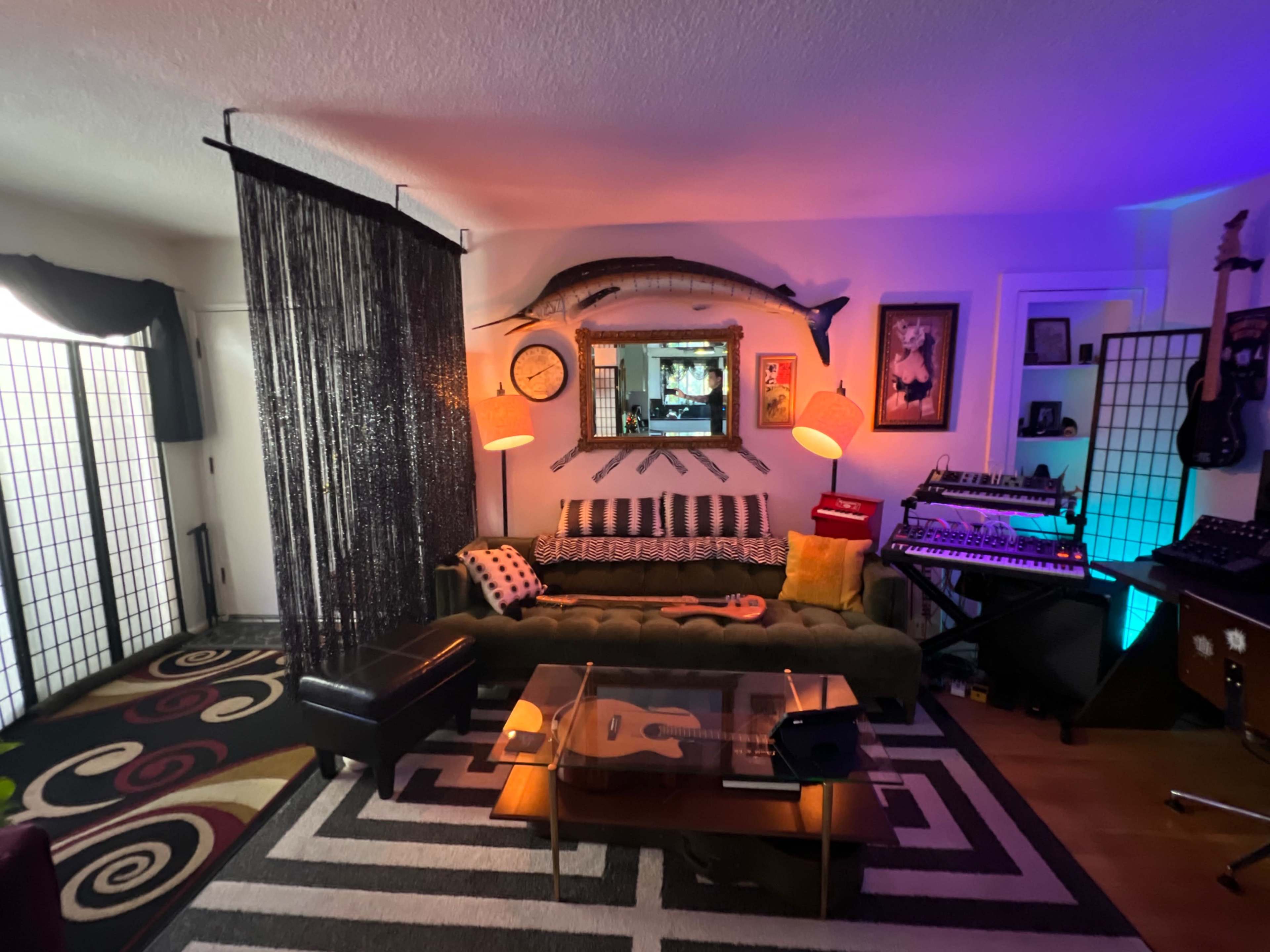 The image shows a stylish living room with a green sofa, striped rug, musical instruments, and decorative screens, illuminated by colorful lighting.