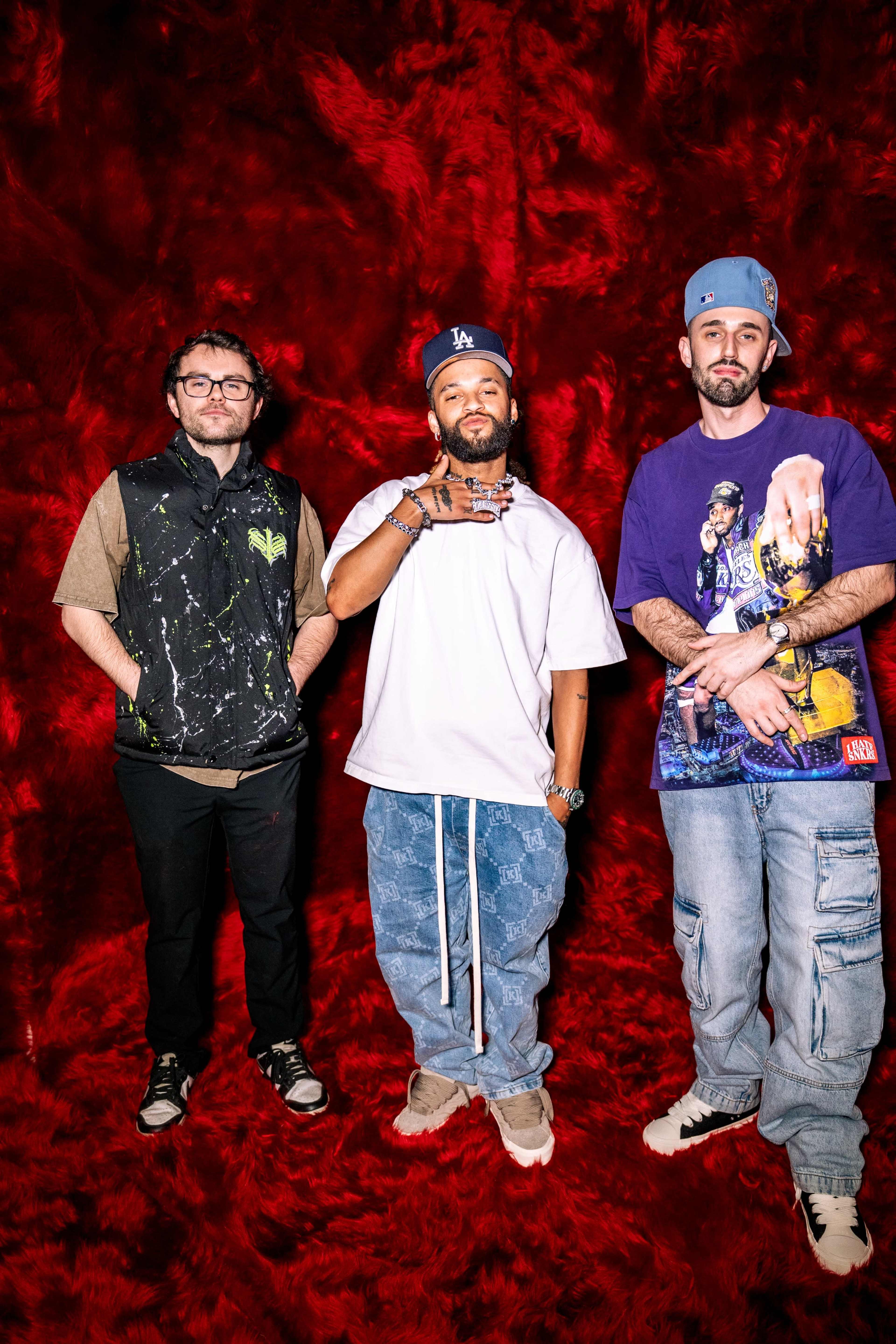 Three men stand in front of a vibrant red backdrop, each wearing distinct casual outfits.