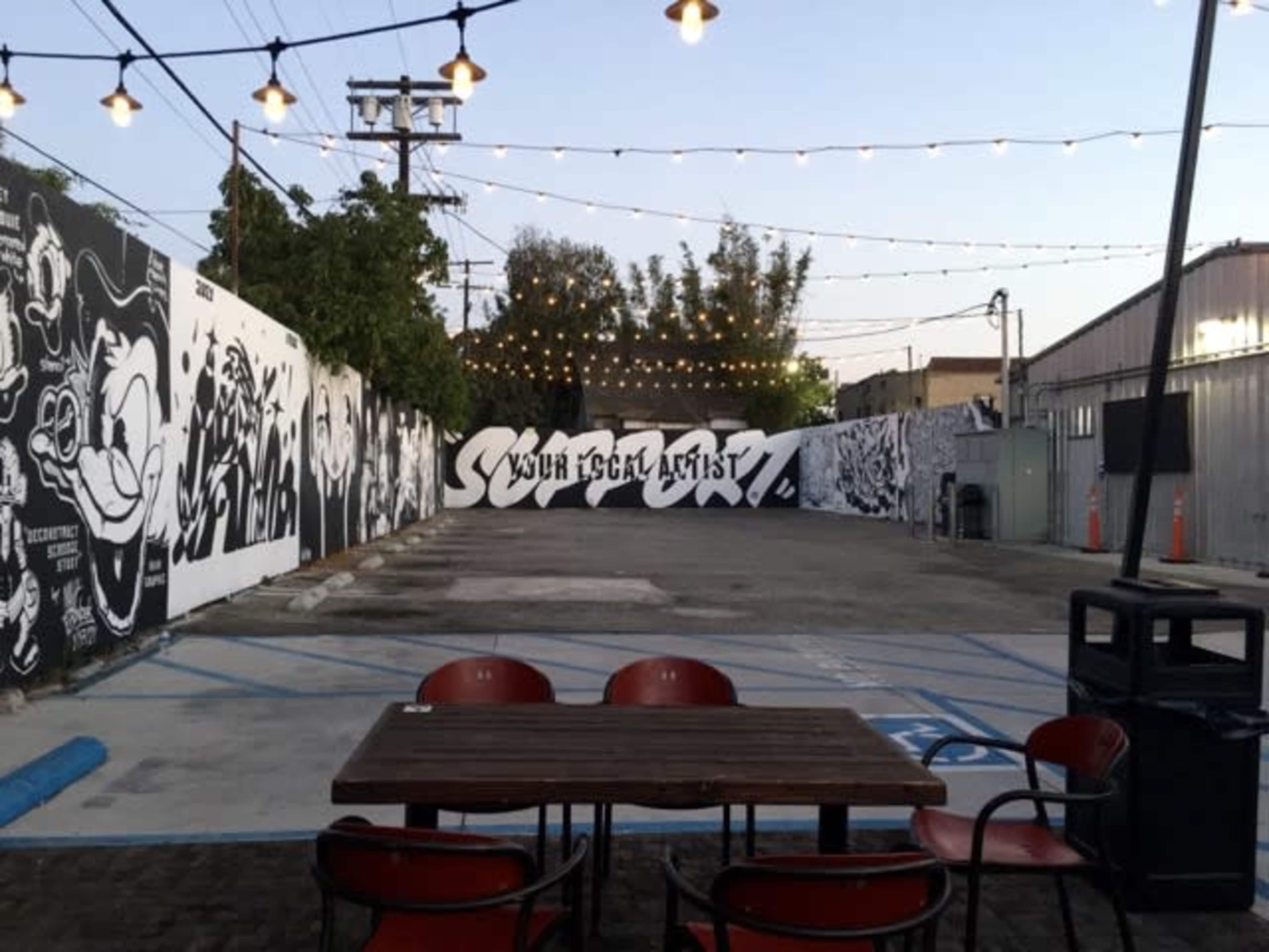 An outdoor space with a table and chairs, surrounded by murals on the walls and string lights overhead.