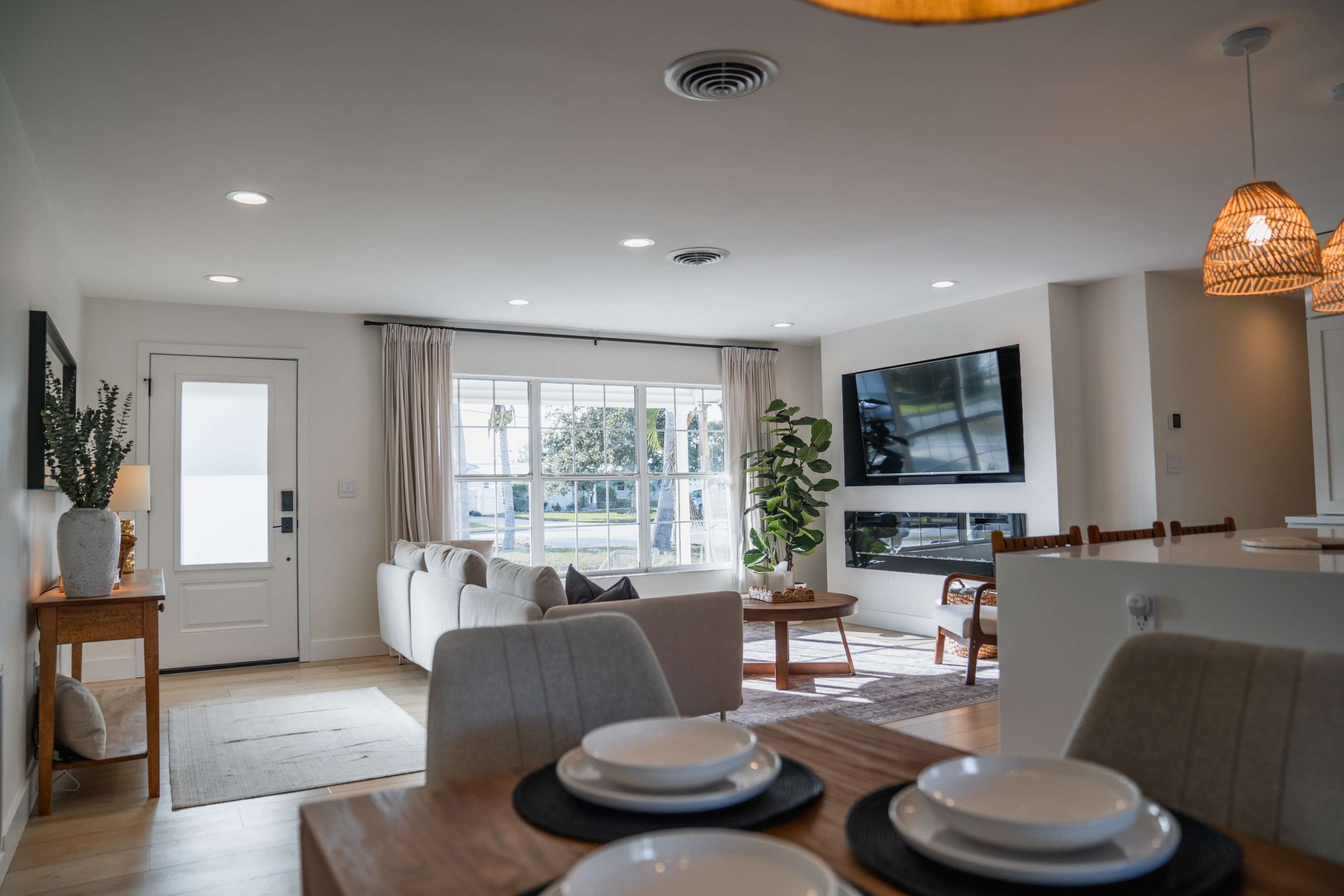 The image shows a modern living room with a large window, a sofa facing a wall-mounted television, a dining area with a wooden table, and natural light illuminating the space.