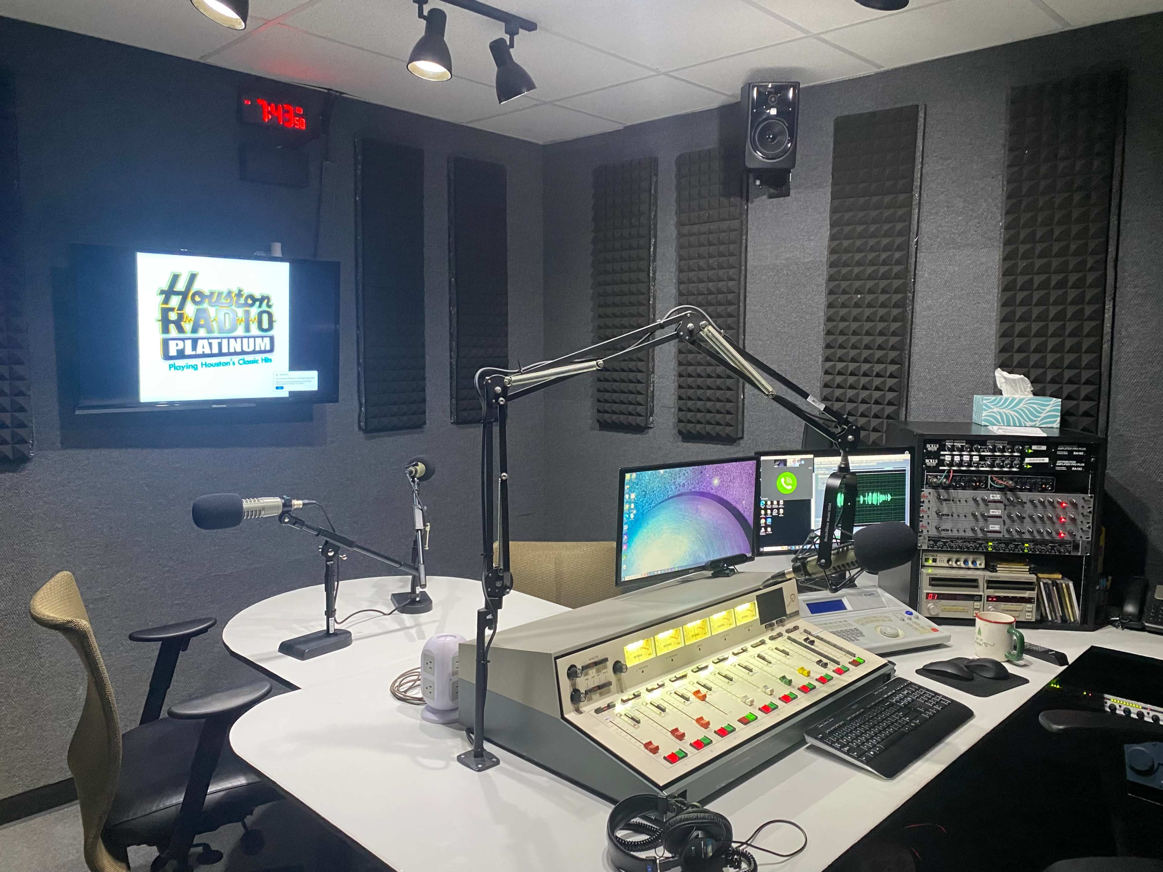 A radio studio featuring a broadcast console, computer monitors, microphones, and soundproof walls, with a screen displaying "Houston Radio Platinum."