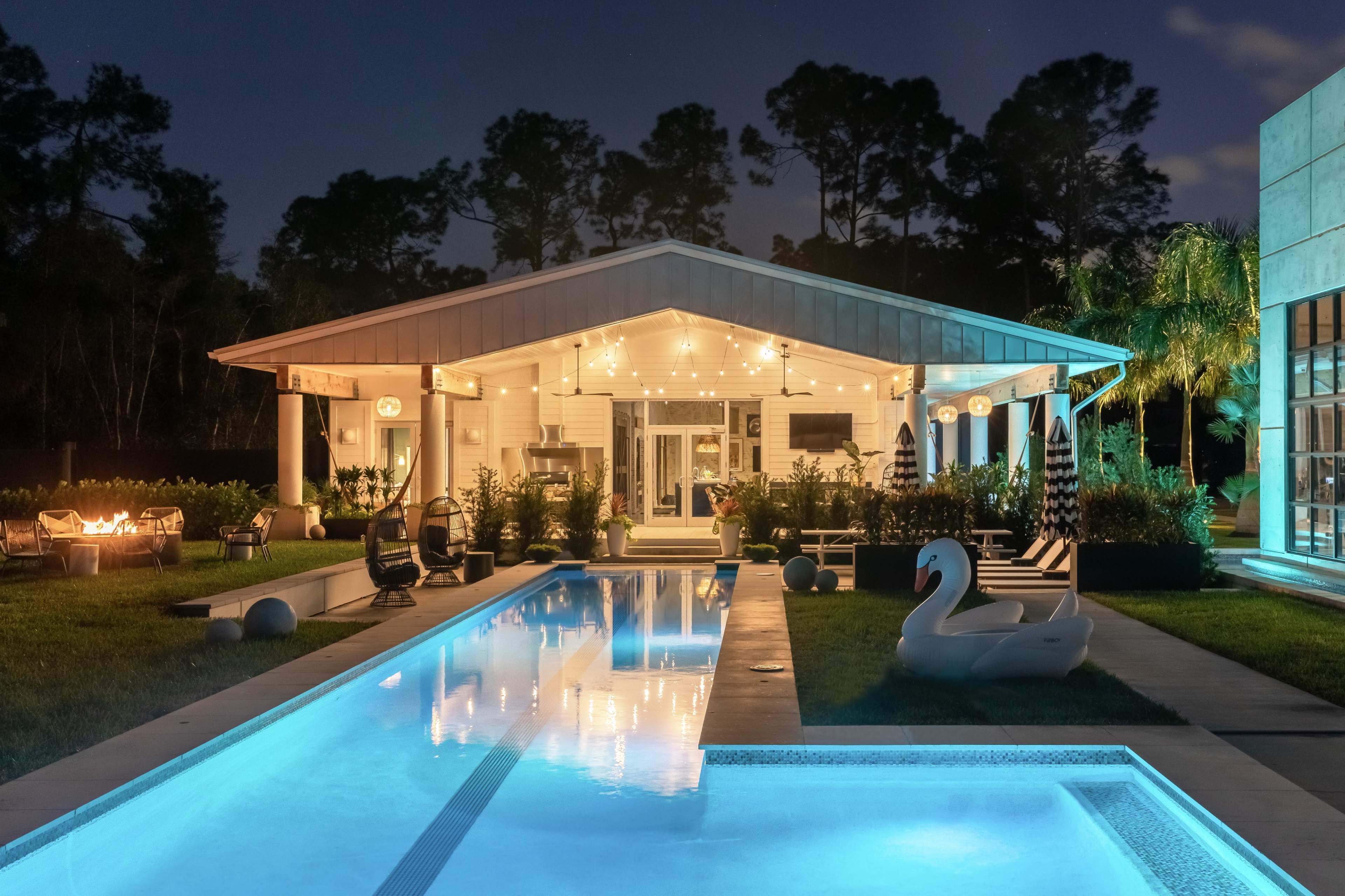 A modern house with outdoor lighting and a pool is set among trees at night, featuring lounge chairs and a decorative swan-shaped float.