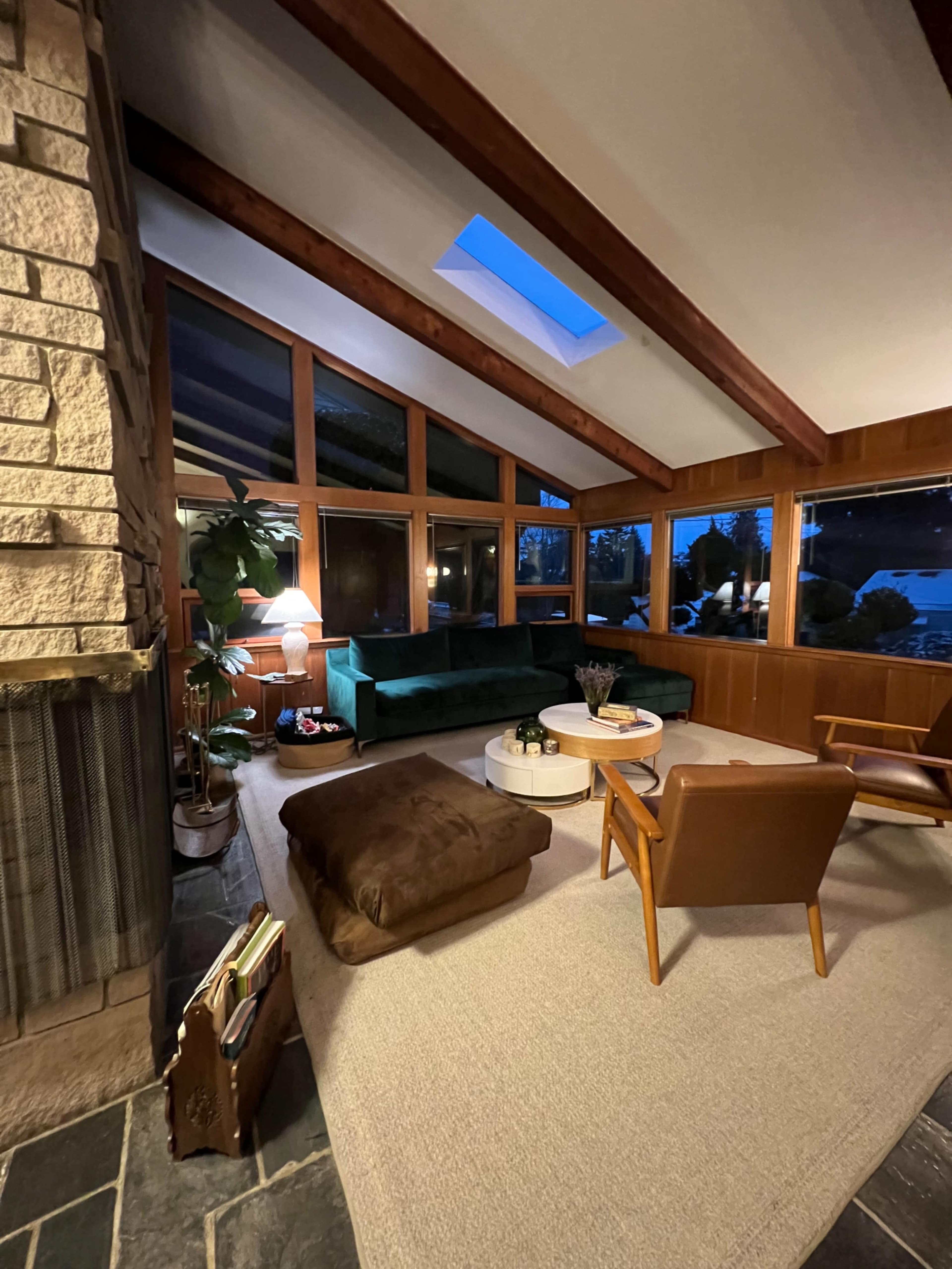 A cozy living room with a stone fireplace, large windows, a green couch, a round coffee table, and a chair, all set on a neutral carpet.
