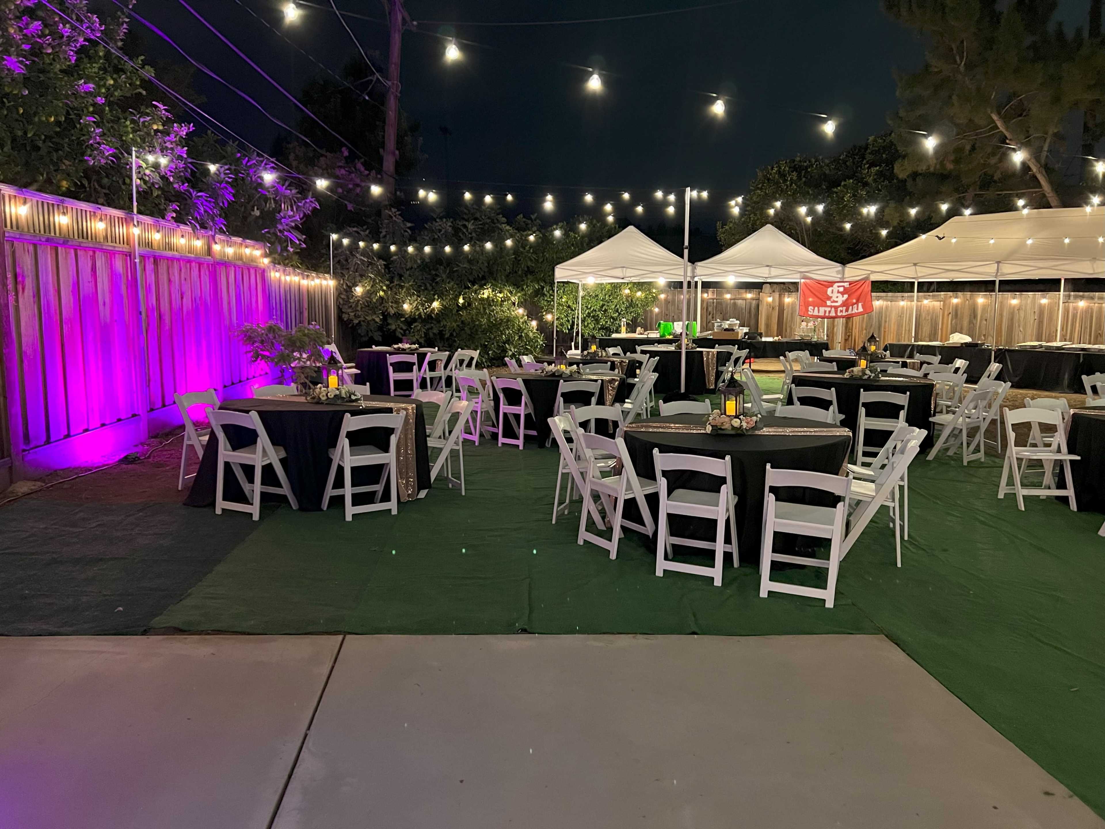 The scene shows a landscaped outdoor event space with tables set for dining, string lights overhead, and canopies in the background.