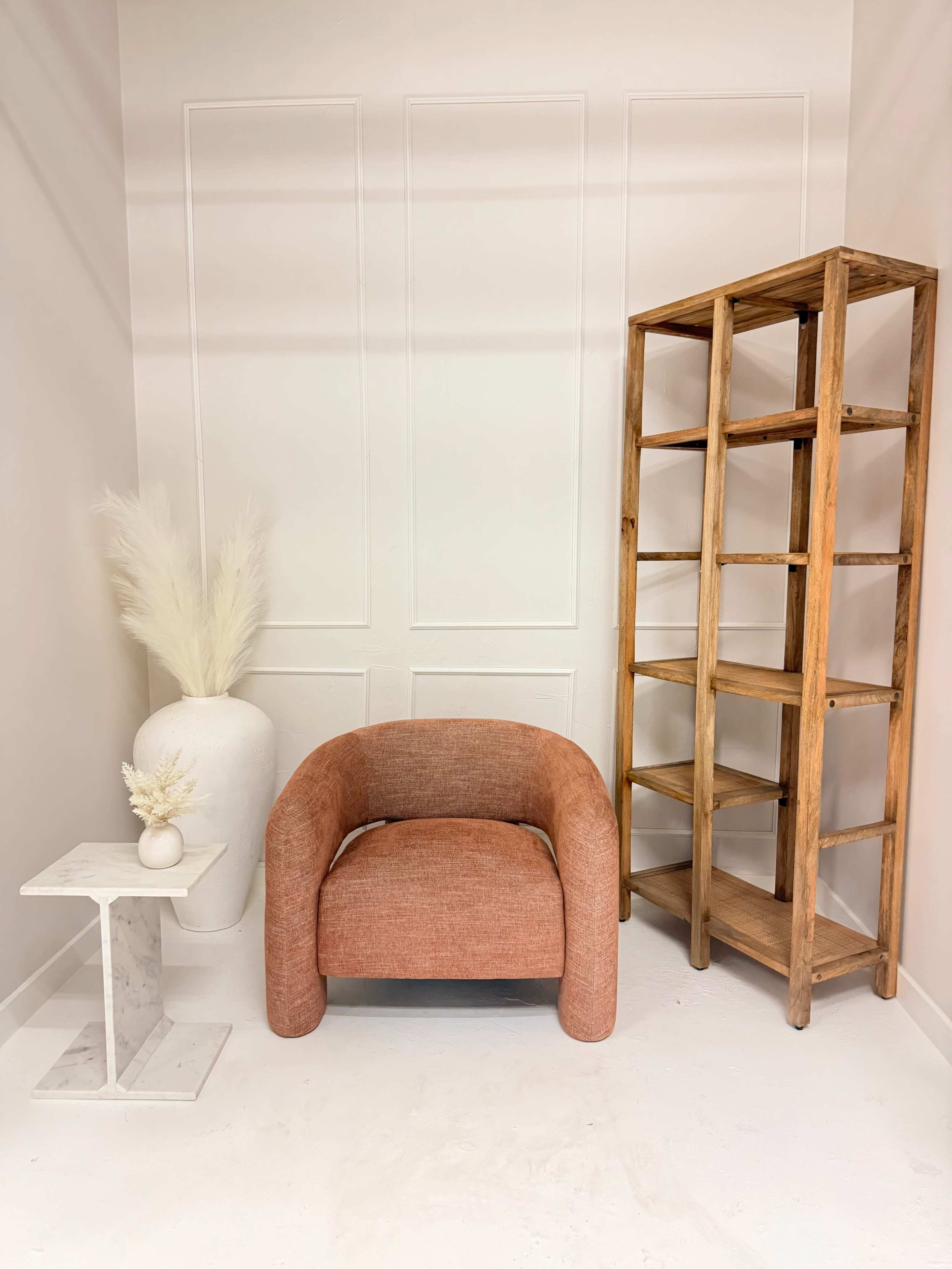 The image features a cozy space with a rounded, pink armchair next to a wooden bookshelf and a marble side table, complemented by a large white vase with dried plants.