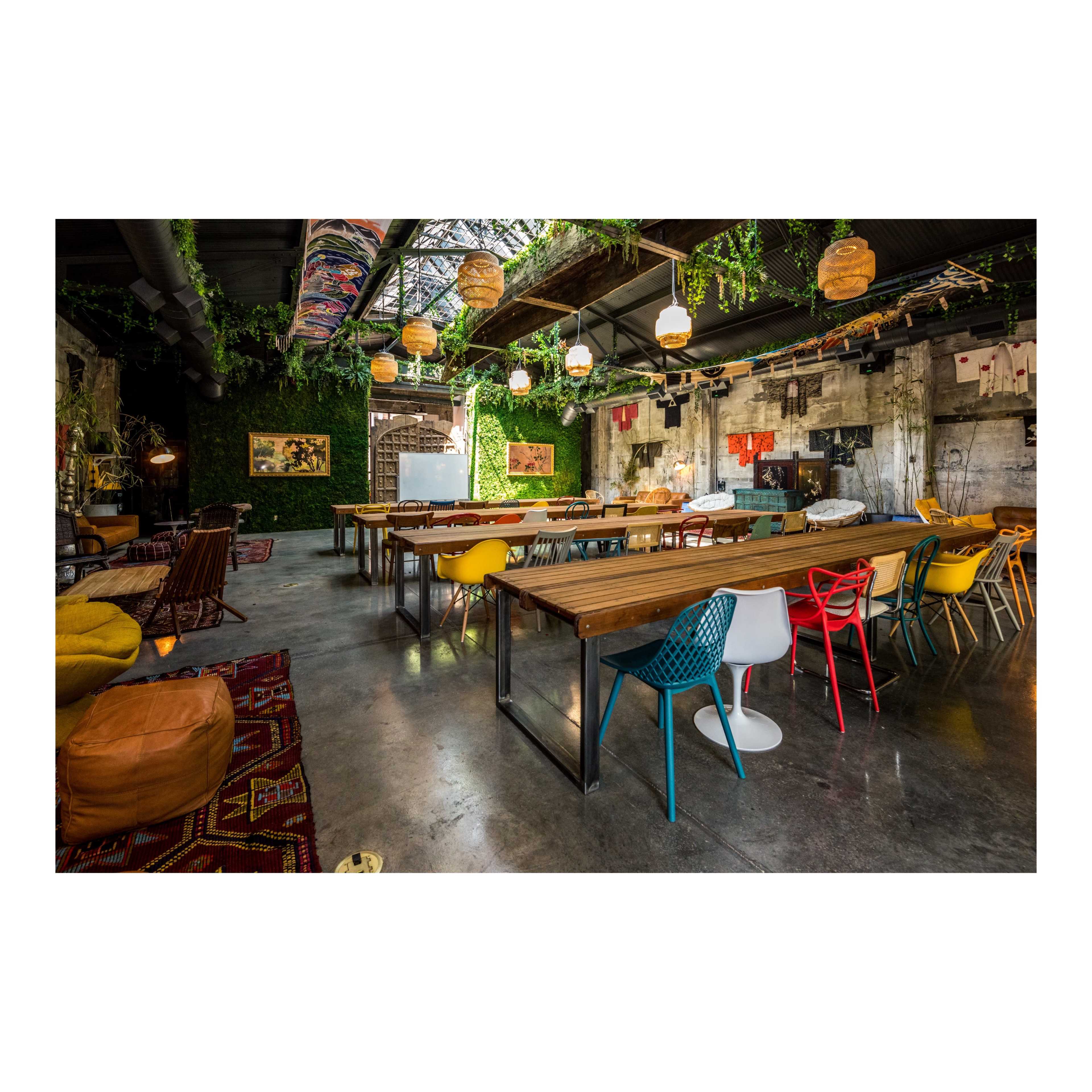 A spacious, industrial-style room with long wooden tables, colorful chairs, and greenery decorating the walls and ceiling.