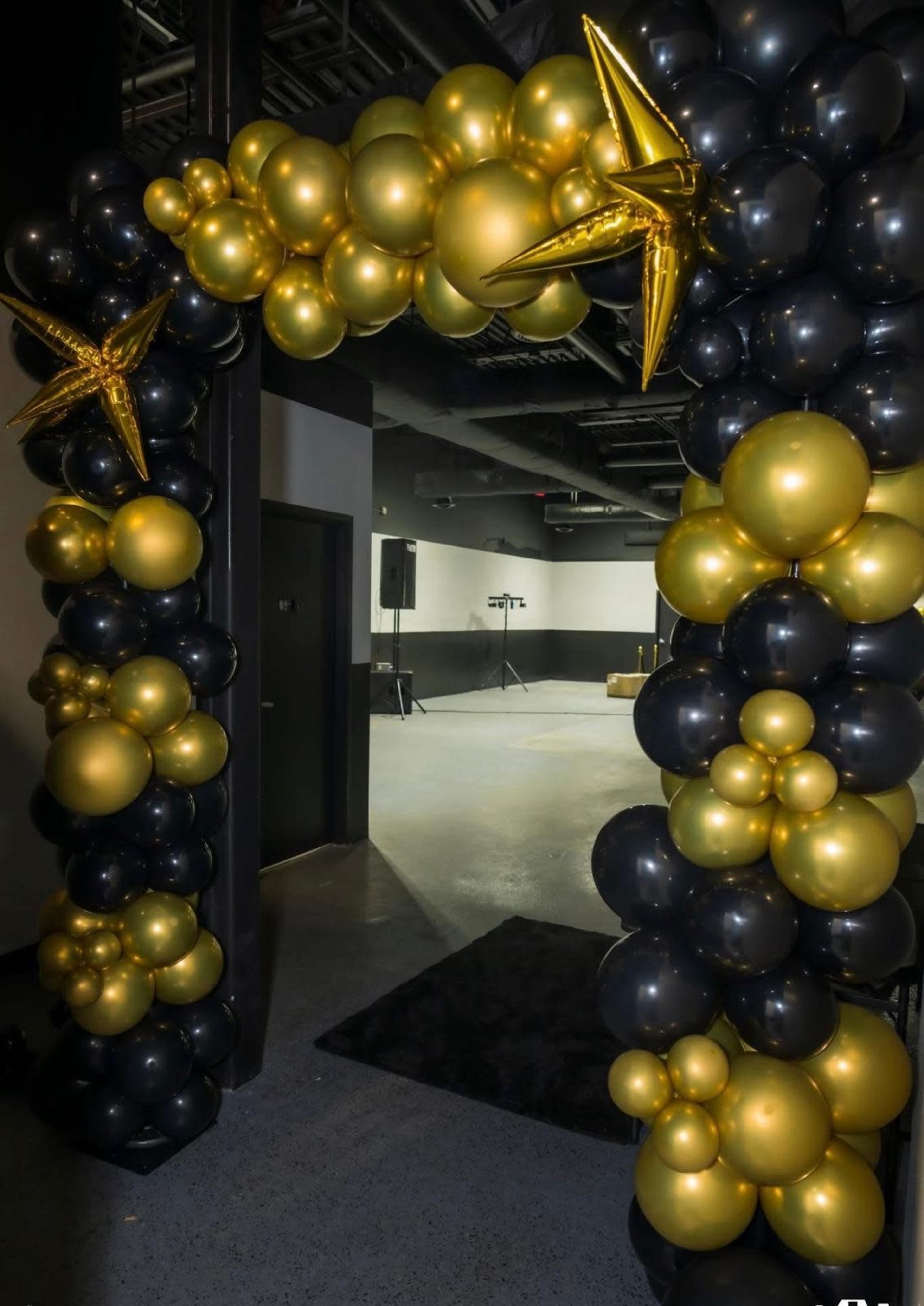 A balloon arch decorated with gold and black balloons frames an entrance to a spacious venue.