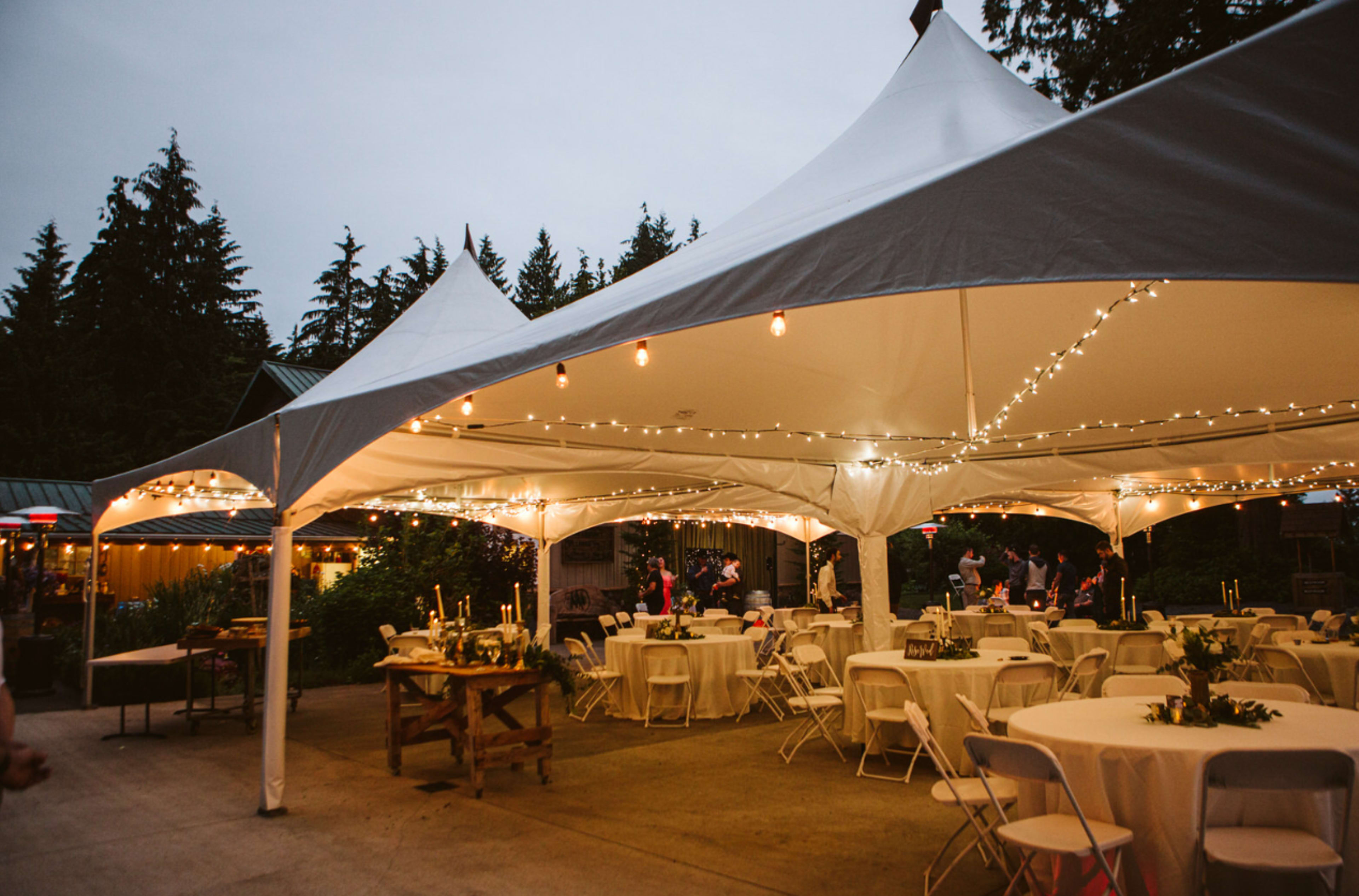 A large outdoor tent is set up with round tables, chairs, and string lights, surrounded by trees and people mingling in the evening.