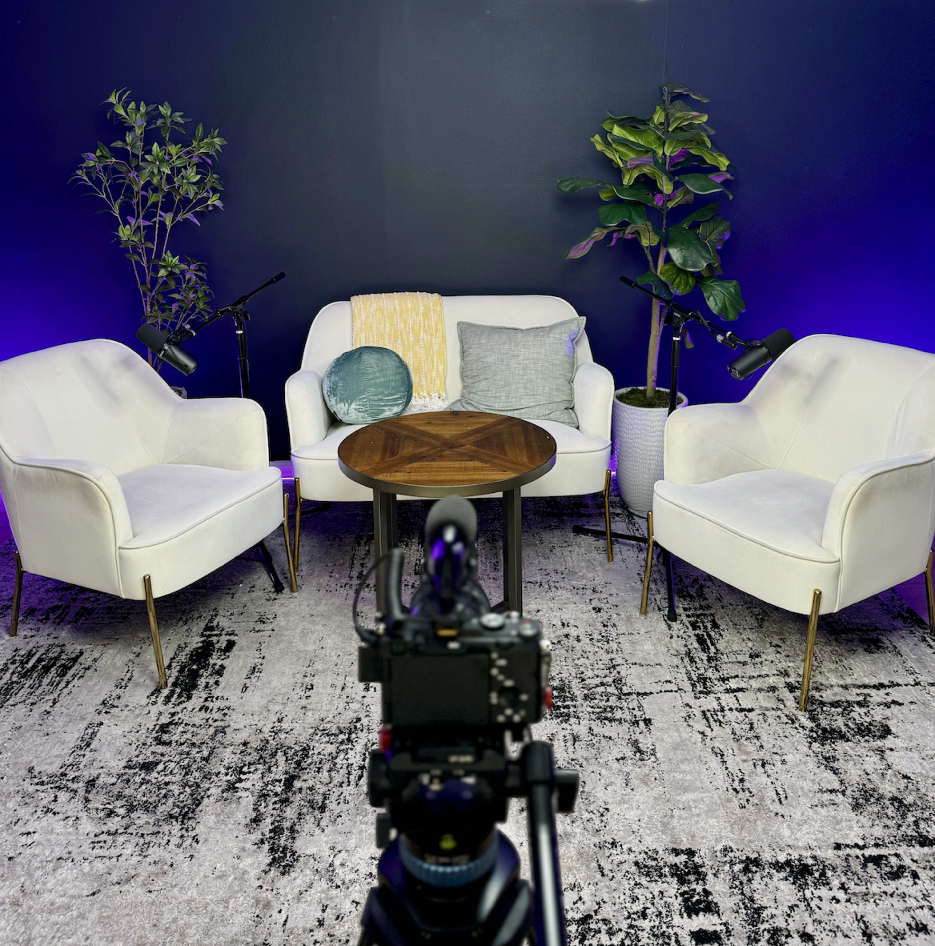 A modern seating area with two white chairs and a round wooden table, surrounded by potted plants against a dark wall.